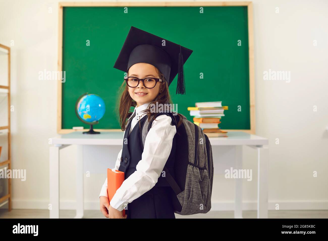 Intelligent little girl in graduation mortar hat posing at classroom. Smart child with backpack on first day of school Stock Photo