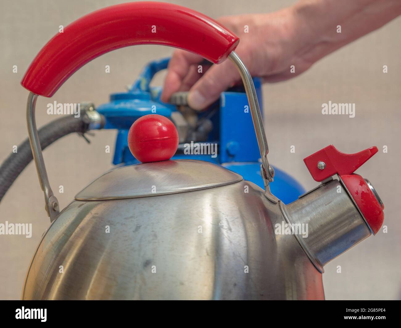 A steel rounded kettle in sharp focus in the foreground, then behind, a man’s fingers loosening the tap on an old blue painted, portable gas bottle. Stock Photo