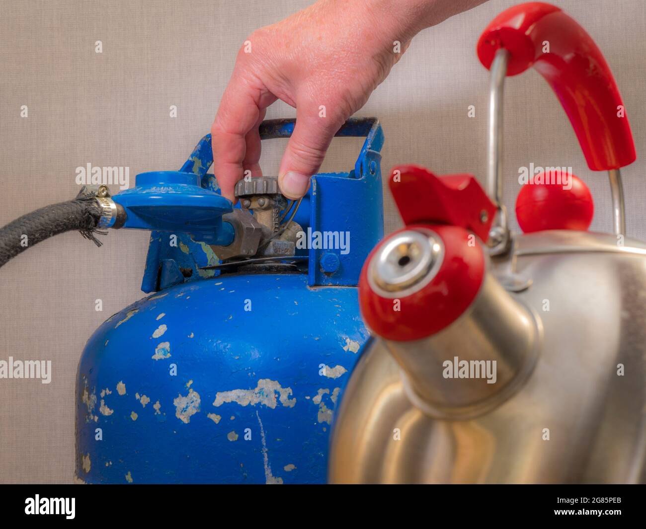 A steel rounded kettle blurred in the foreground, then behind in focus, a man’s fingers loosening the tap on an old blue painted, portable gas bottle. Stock Photo