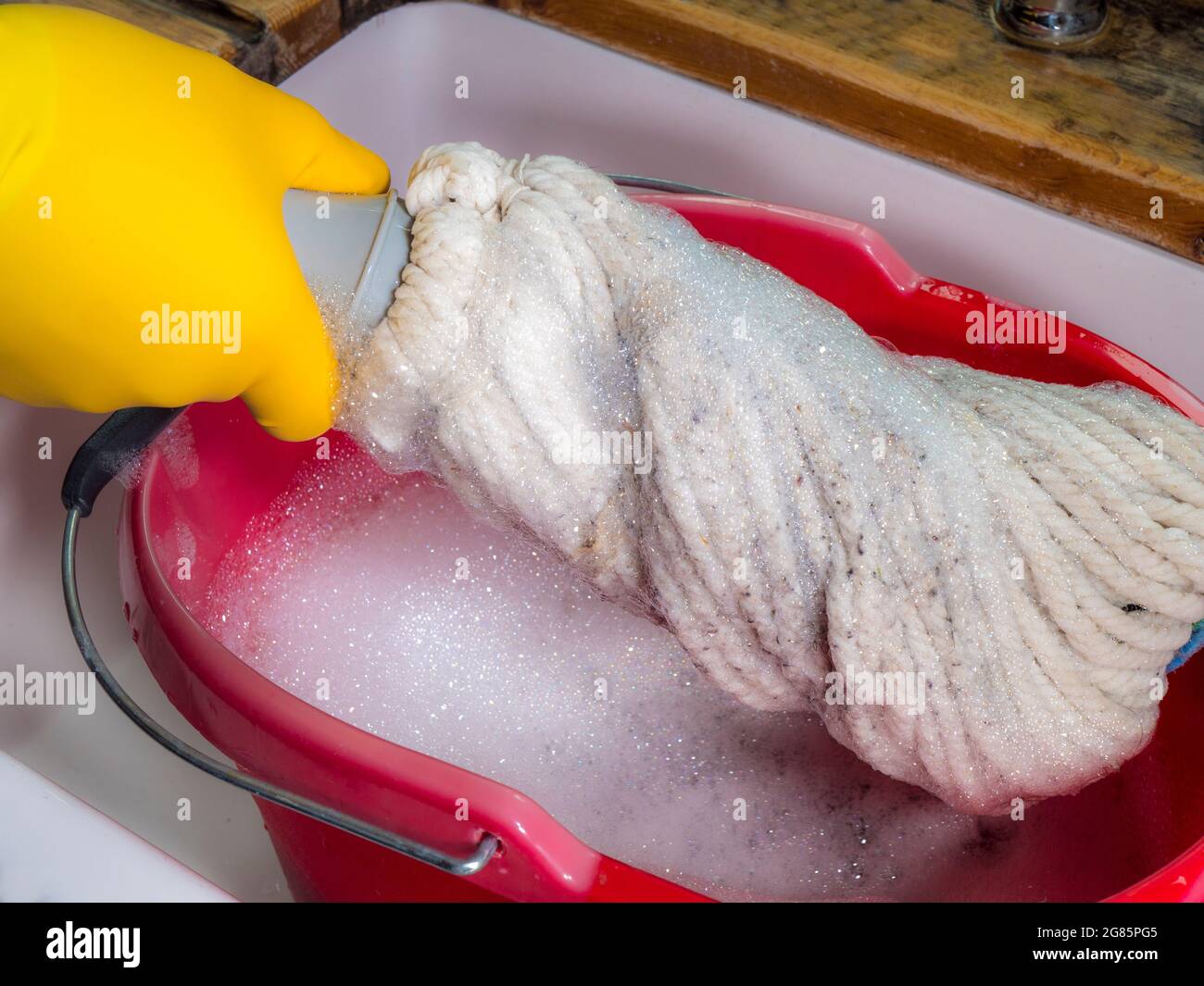 Closeup POV overhead shot of a hand in a yellow rubber glove twisting a mop over a red plastic bucket in a sink, to squeeze out soapy water. Stock Photo