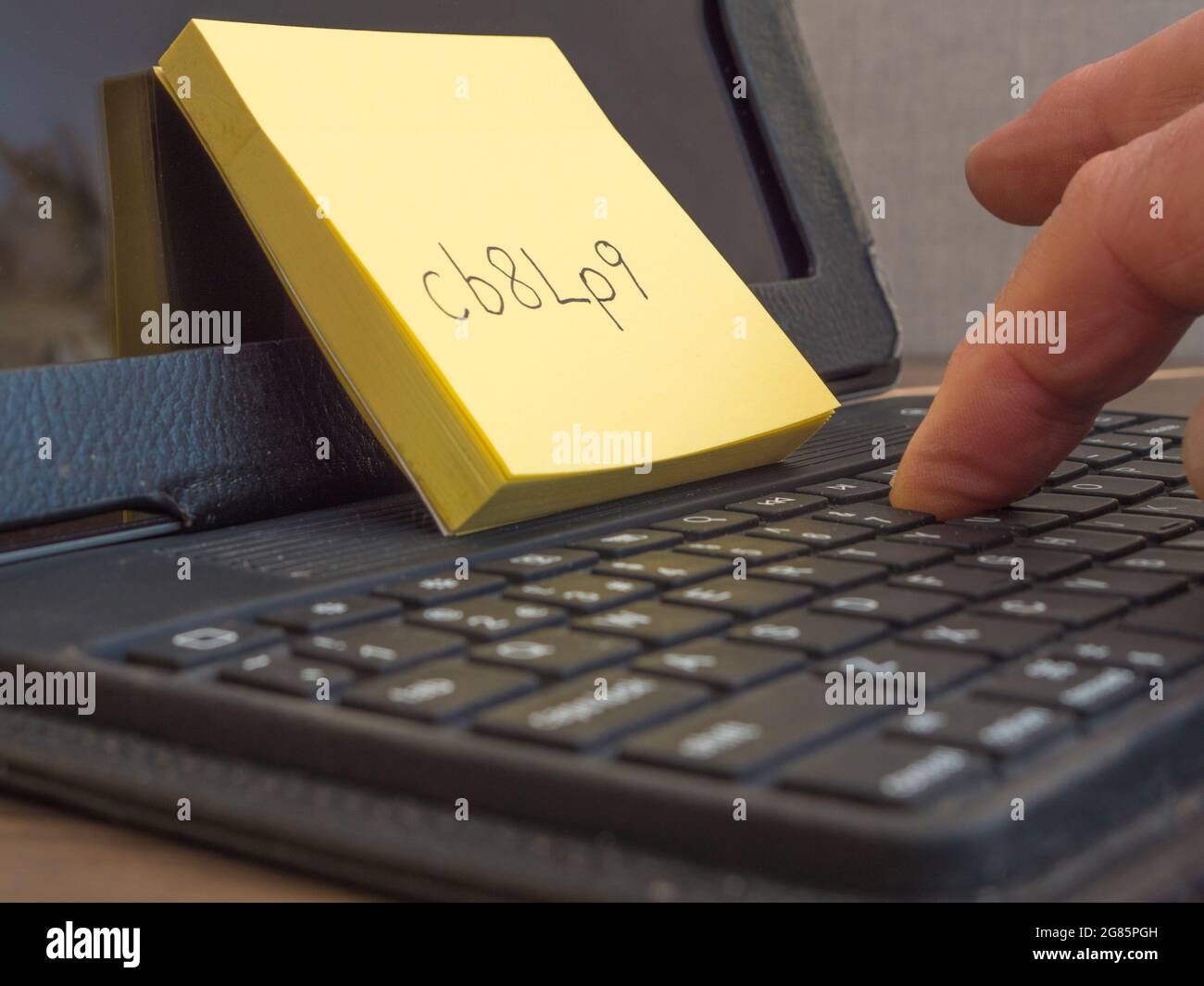 Closeup of a man’s finger typing on a portable keyboard, an unfamiliar computer password, written on a notepad leaning against a tablet screen. Stock Photo