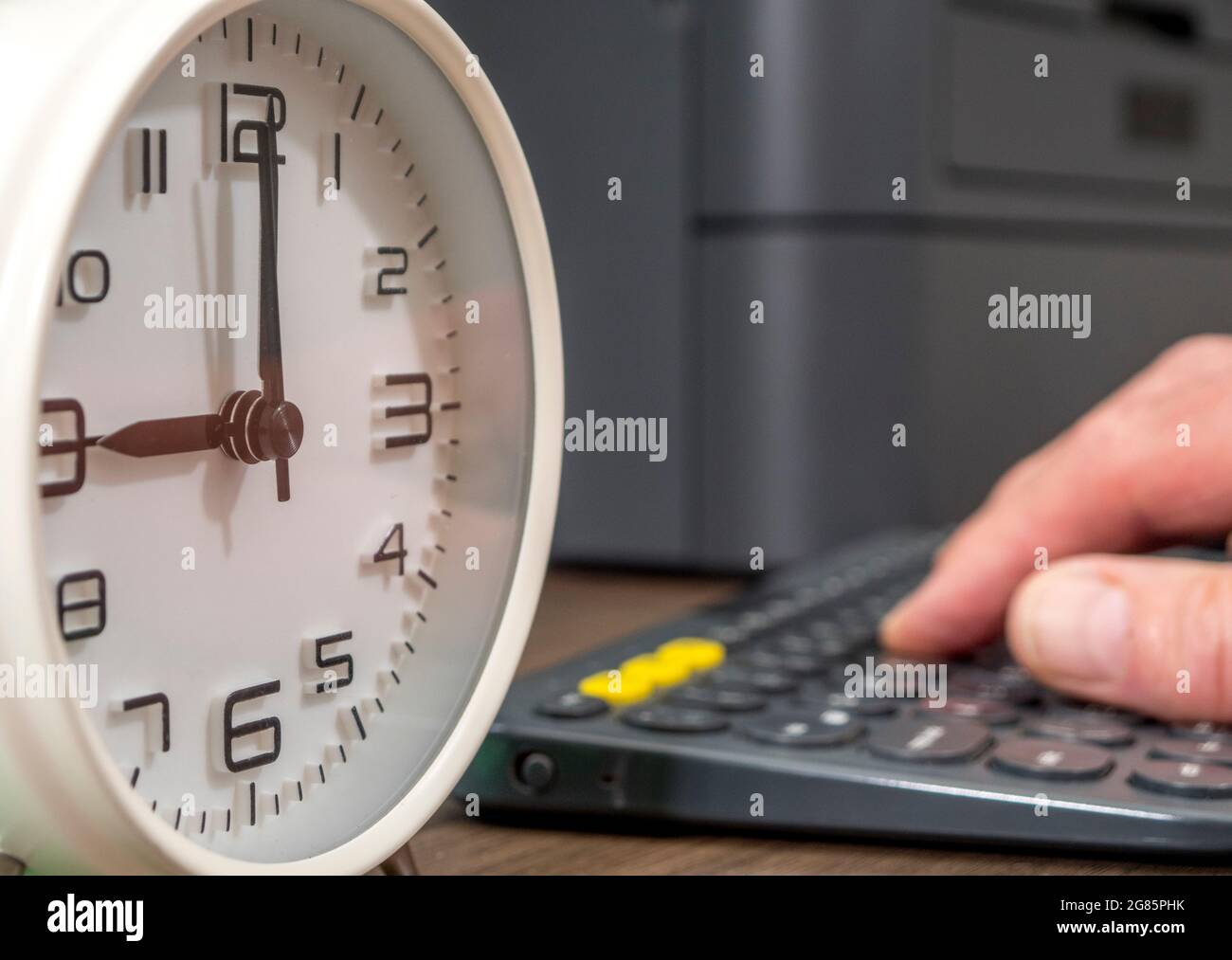 Closeup of defocused man’s fingers using a computer keyboard on a desk, with focus on a clock face showing 9 o’clock. Concept of starting office work. Stock Photo