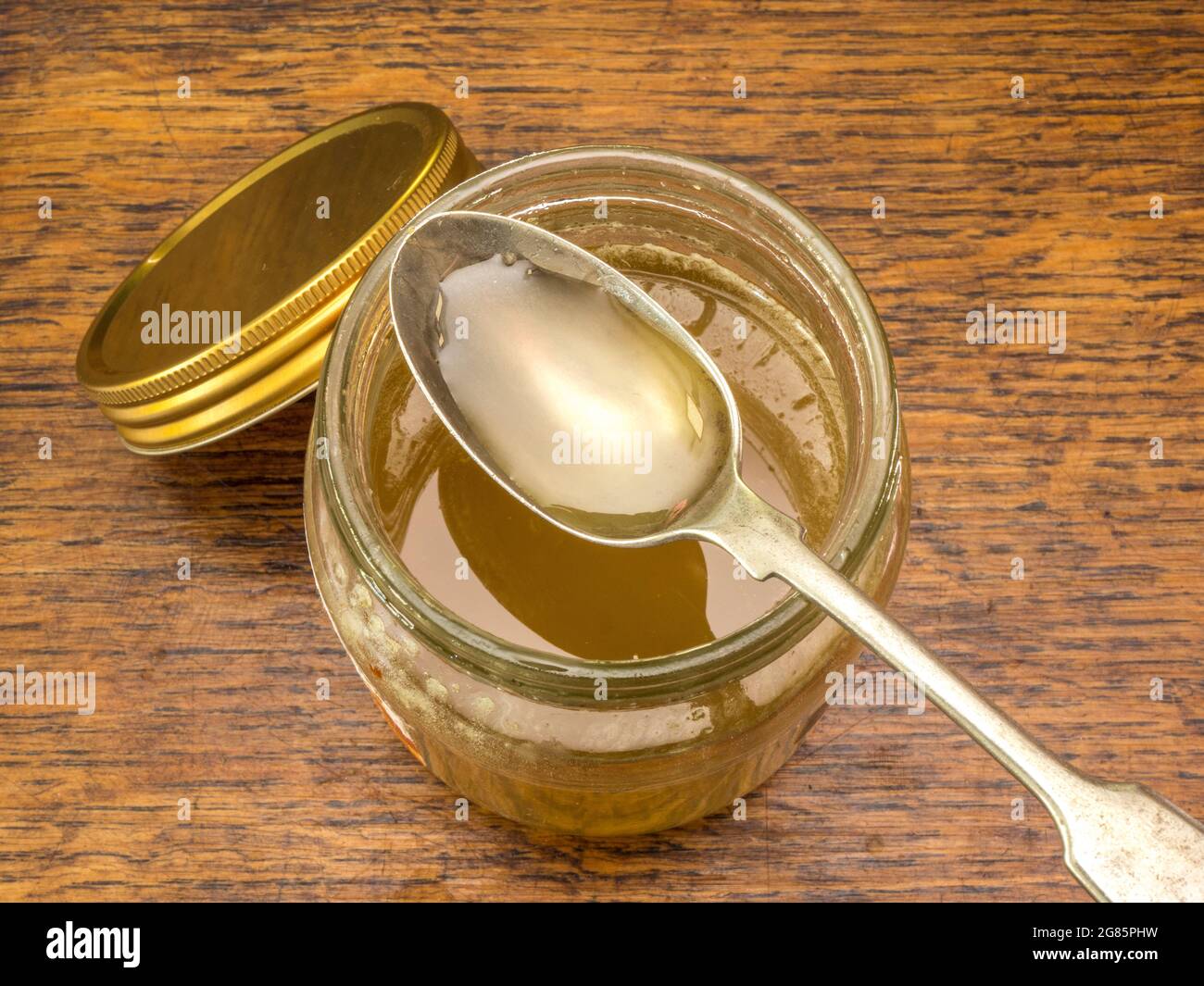 Closeup overhead view of a spoonful of thick pure bee honey resting on the top of an open product jar, standing on a wooden table. Stock Photo