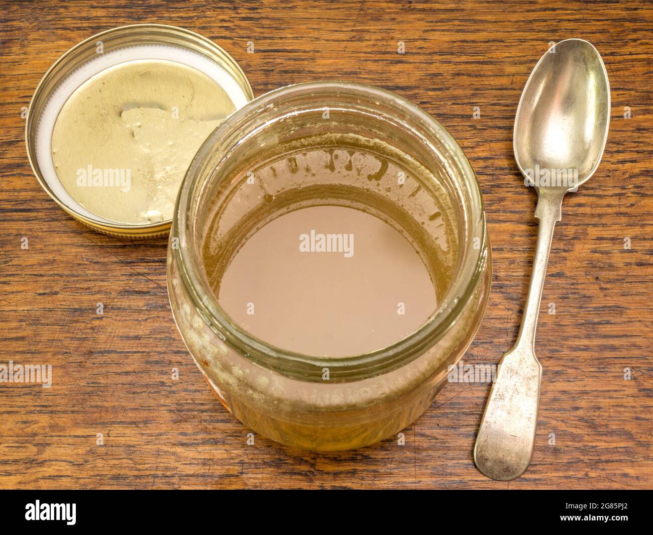 Closeup overhead view of an open product jar of thick pure bee honey, standing on a wooden table, with the lid and a spoon nearby. Stock Photo