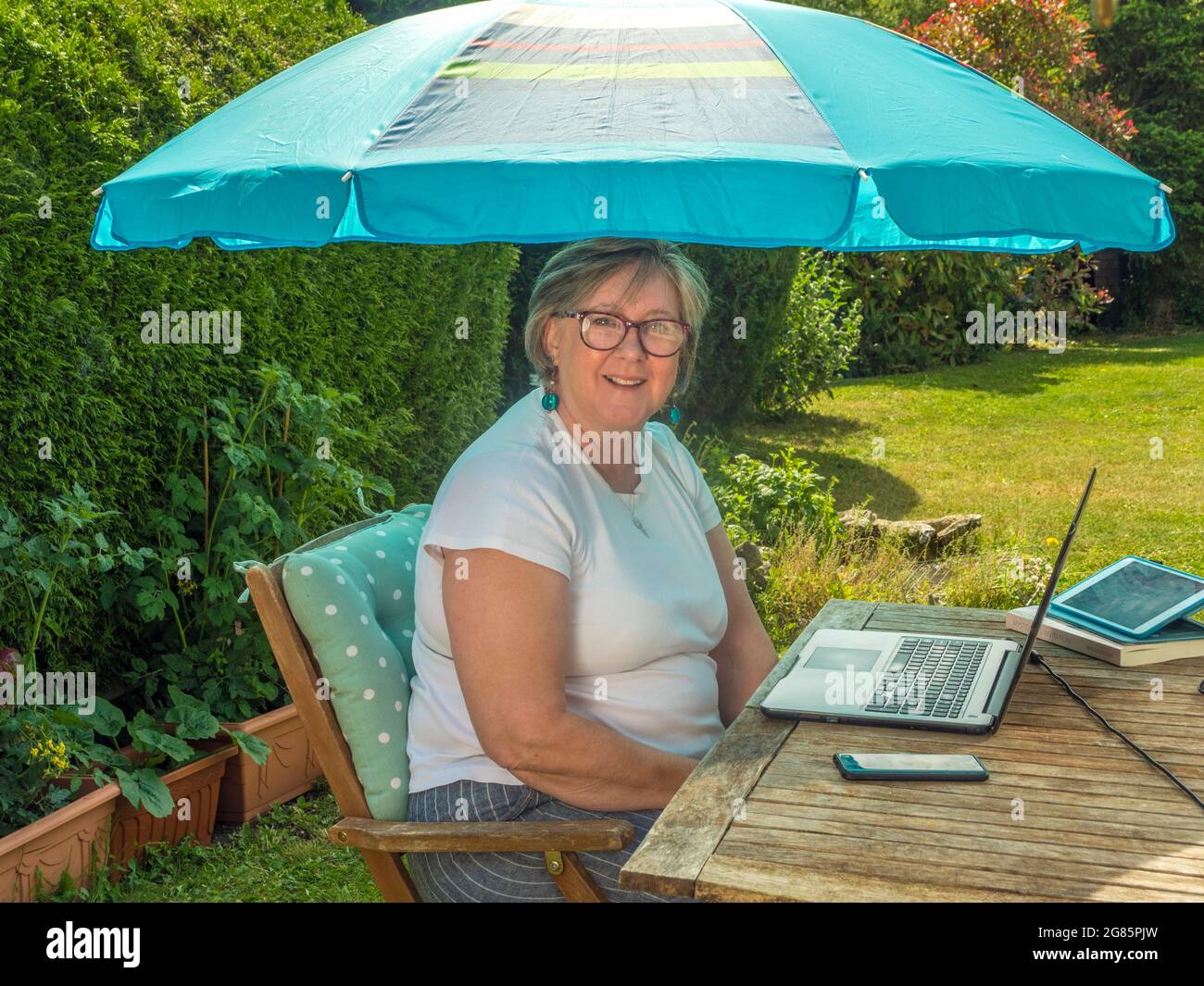 A mature woman sitting in a chair at a table, outside in a sunny garden / yard, under a sunshade, using a laptop computer, facing camera and smiling. Stock Photo