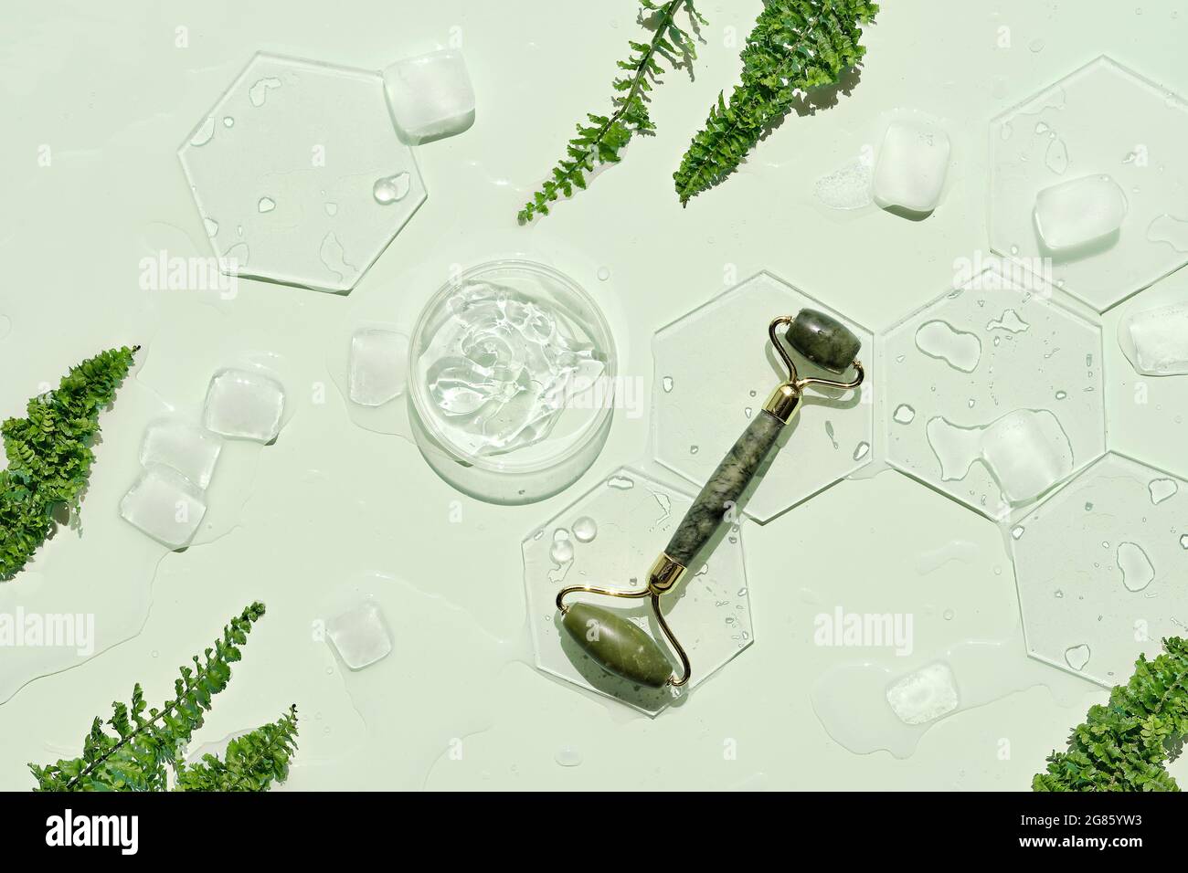 Self made moisturizer, green jade face roller with ice cubes. Exotic fern leaves on mint green background. Minimal flat lay, top view. Facial massage Stock Photo