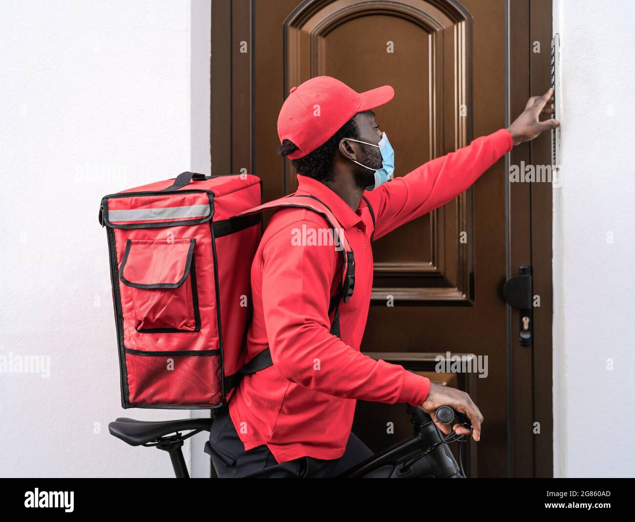 African rider man delivering meal to customers home with electric bicycle while wearing face mask during corona virus outbreak Stock Photo