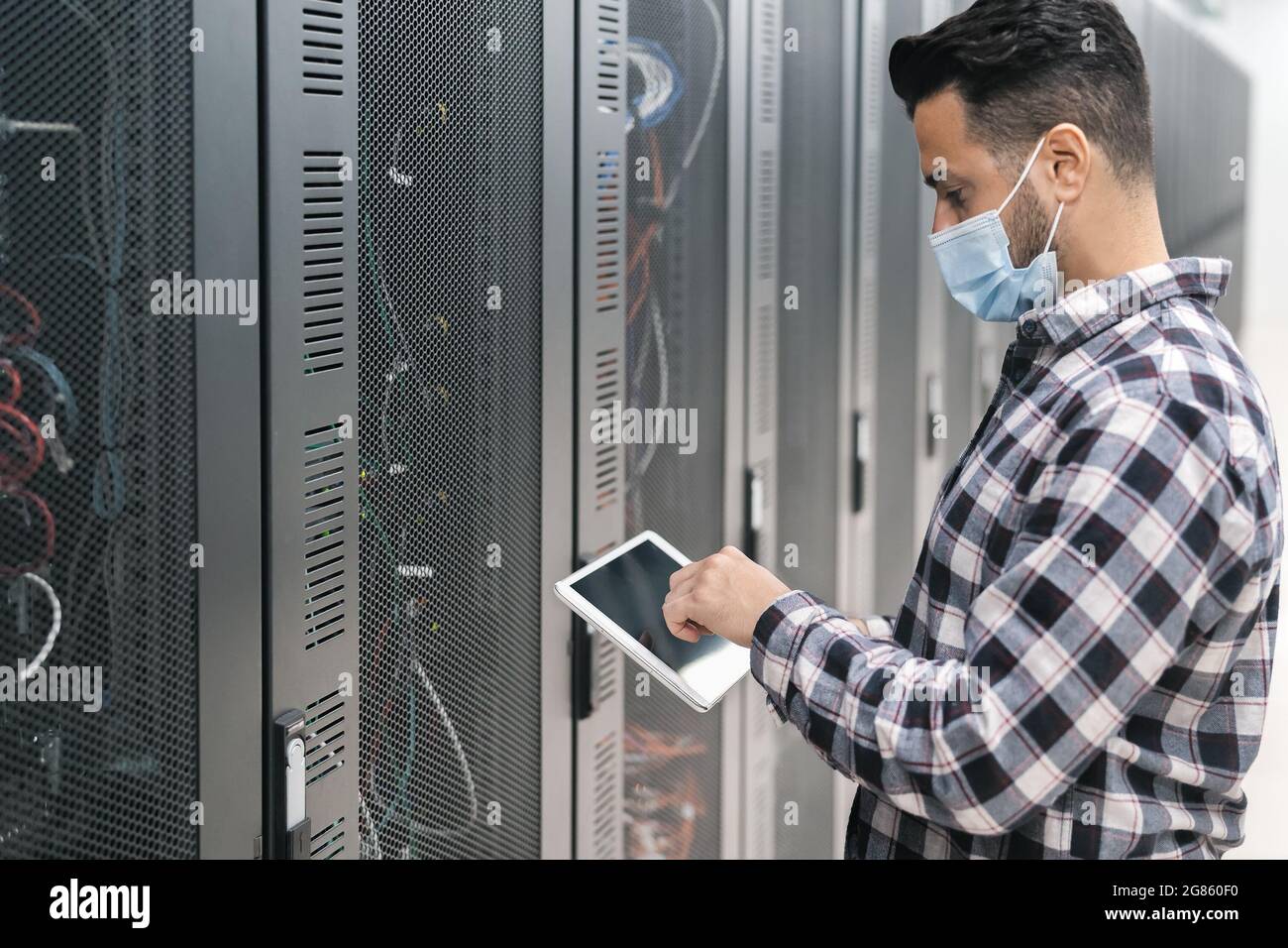 Male informatic engineer working inside server room database while wearing face mask during corona virus outbreak Stock Photo