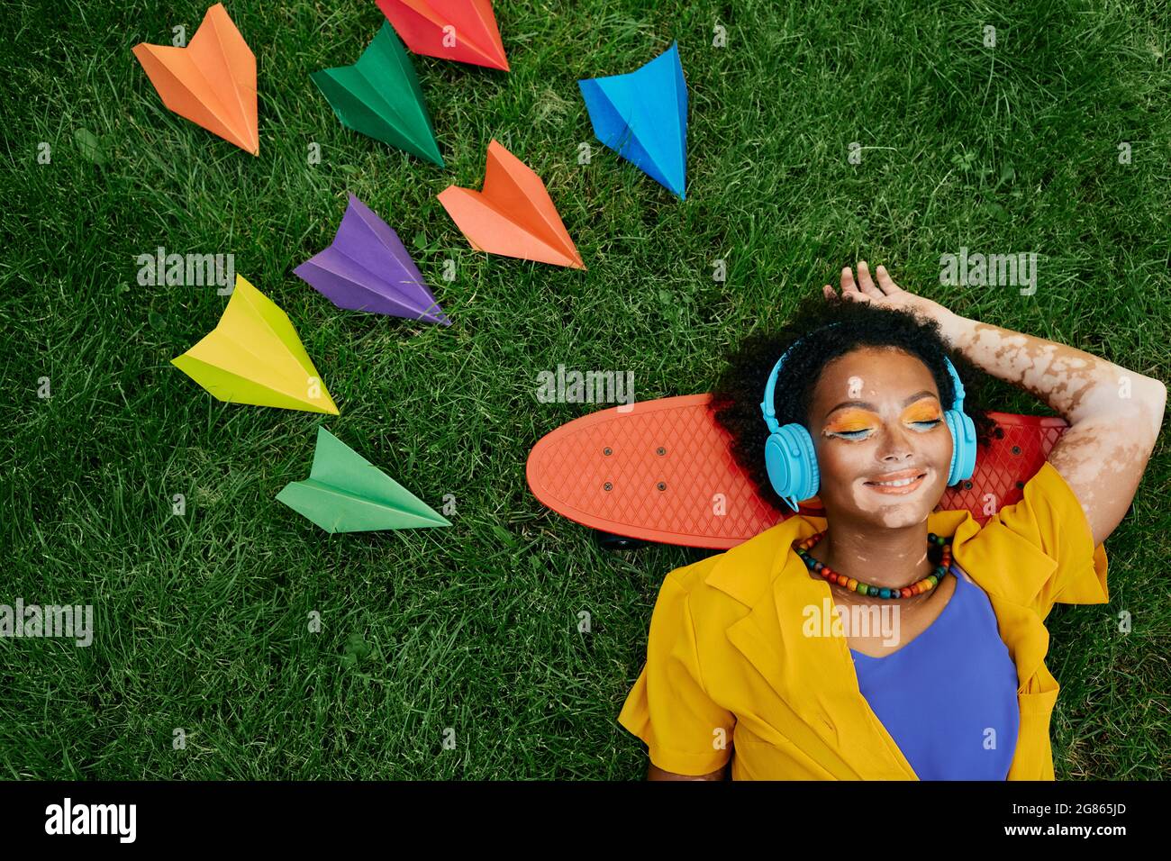Multi-ethnic woman wearing blue headphones listens to music lying on the grass near skateboard with closed eyes and dreams of flying on airplane and t Stock Photo