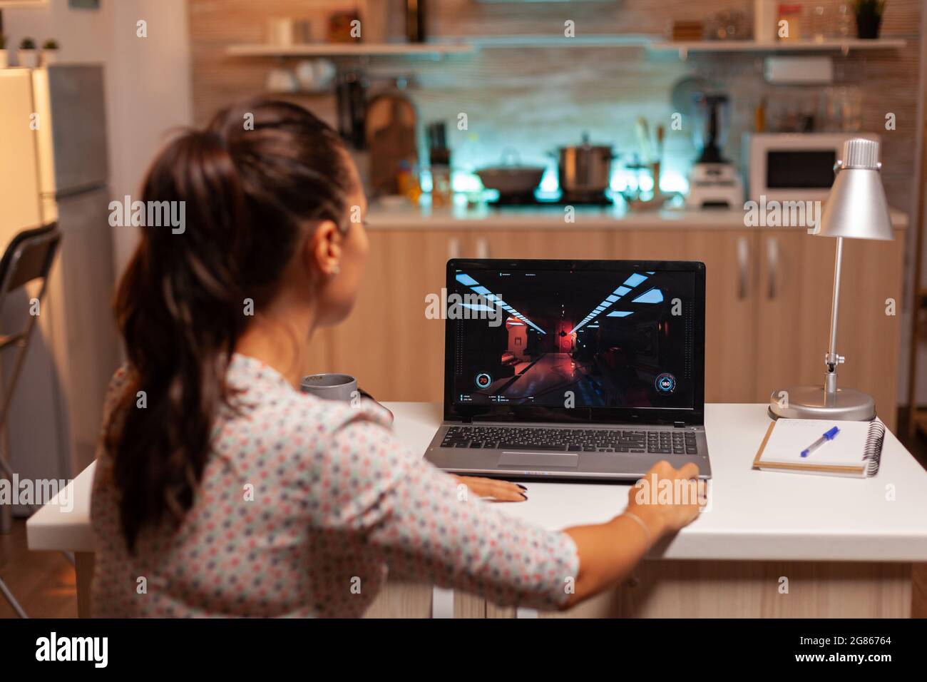 Female game developer working on new project on personal laptop in home kitchen during night time. Professional gamer playing online videop games on her personal computer. Geek cyber e-sport. Stock Photo