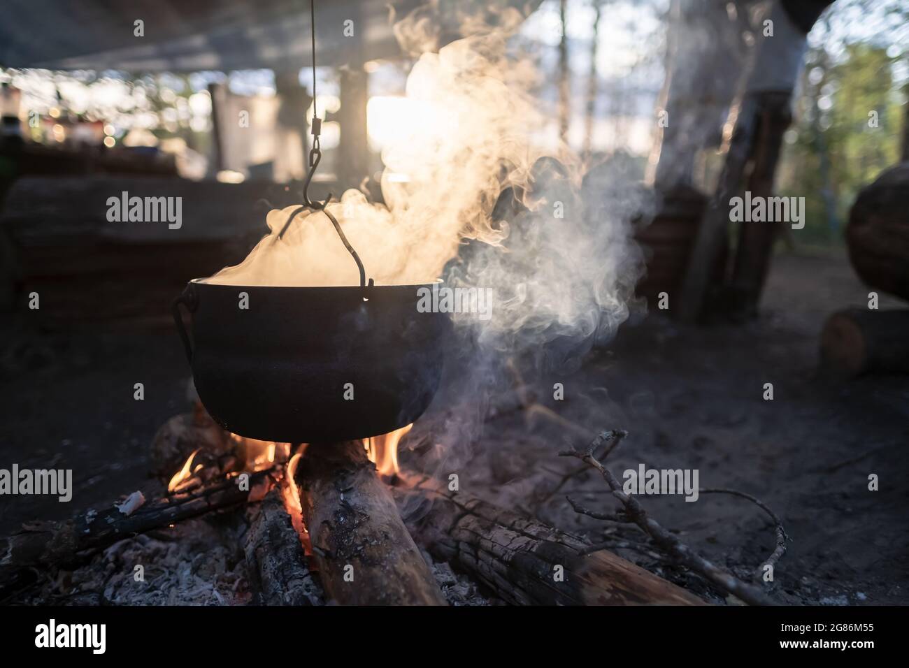 Bowler with escaping steam hangs over a campfire, in which food is prepared, at sunset, against the backdrop of a lake. Stock Photo