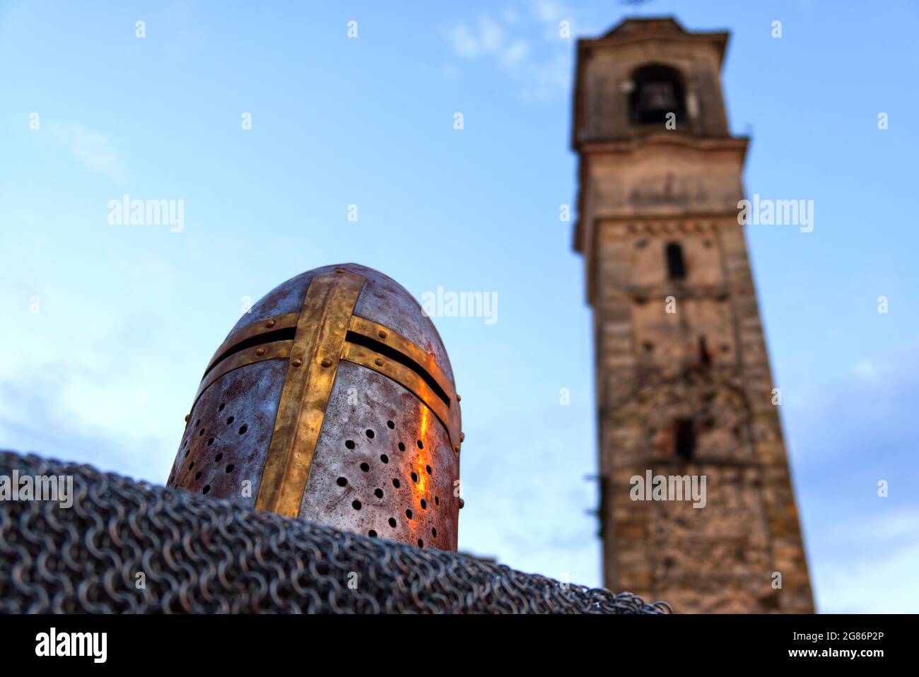 close up of a helmet resting on a chain mail with a Romanesque bell tower in the background Stock Photo