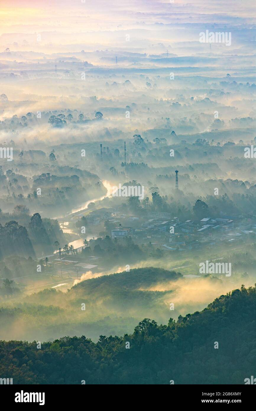 View of sunrise with sun rays illuminated the beautiful panorama of village shrouded in soft fog. Far and several layers of mountain landscape surroun Stock Photo