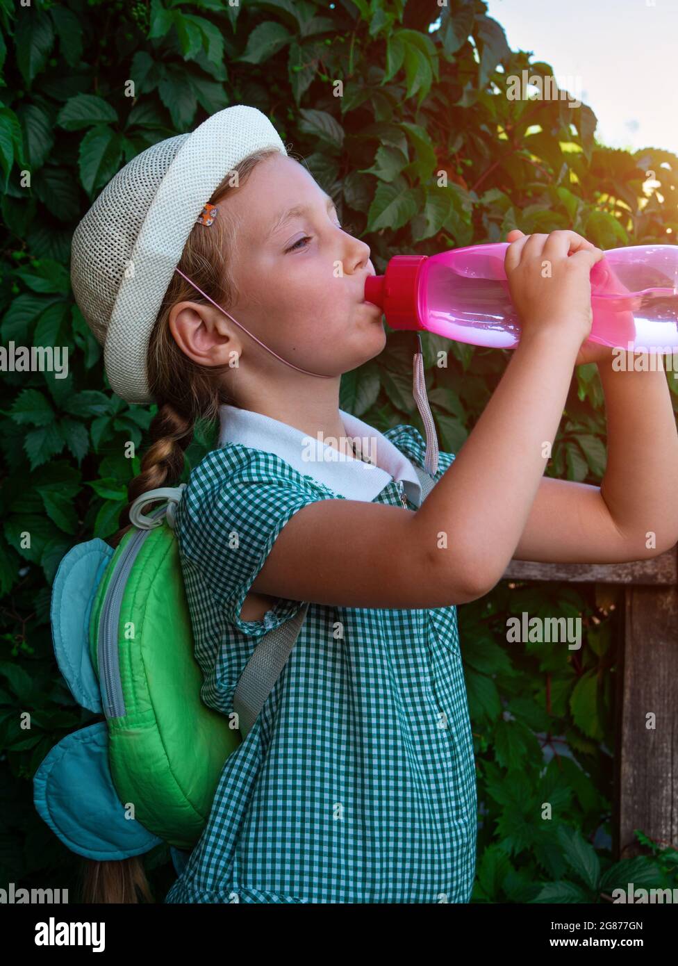 Cute school little girl drinks water from reusable pink bottle outdoor. Child in hat enjoys