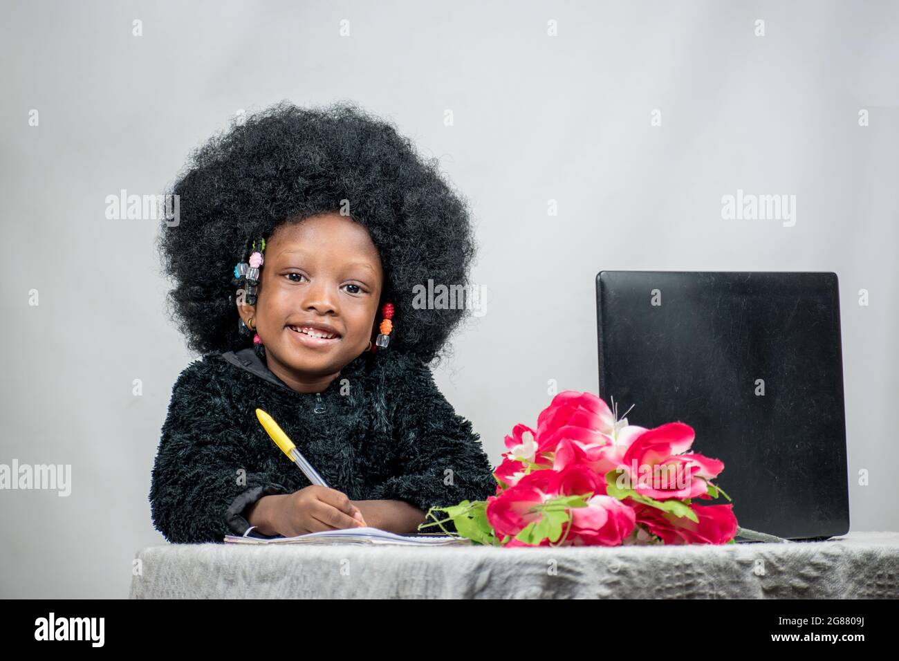 African girl child with afro hair, writing with a pen, studying and researching for excellence in her education with a laptop, flower on her table Stock Photo