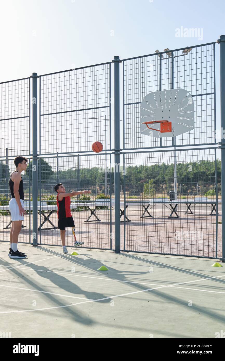 Basketball trainer showing how to shoot basketball to a child with a leg prosthesis. Coach training a kid. Stock Photo