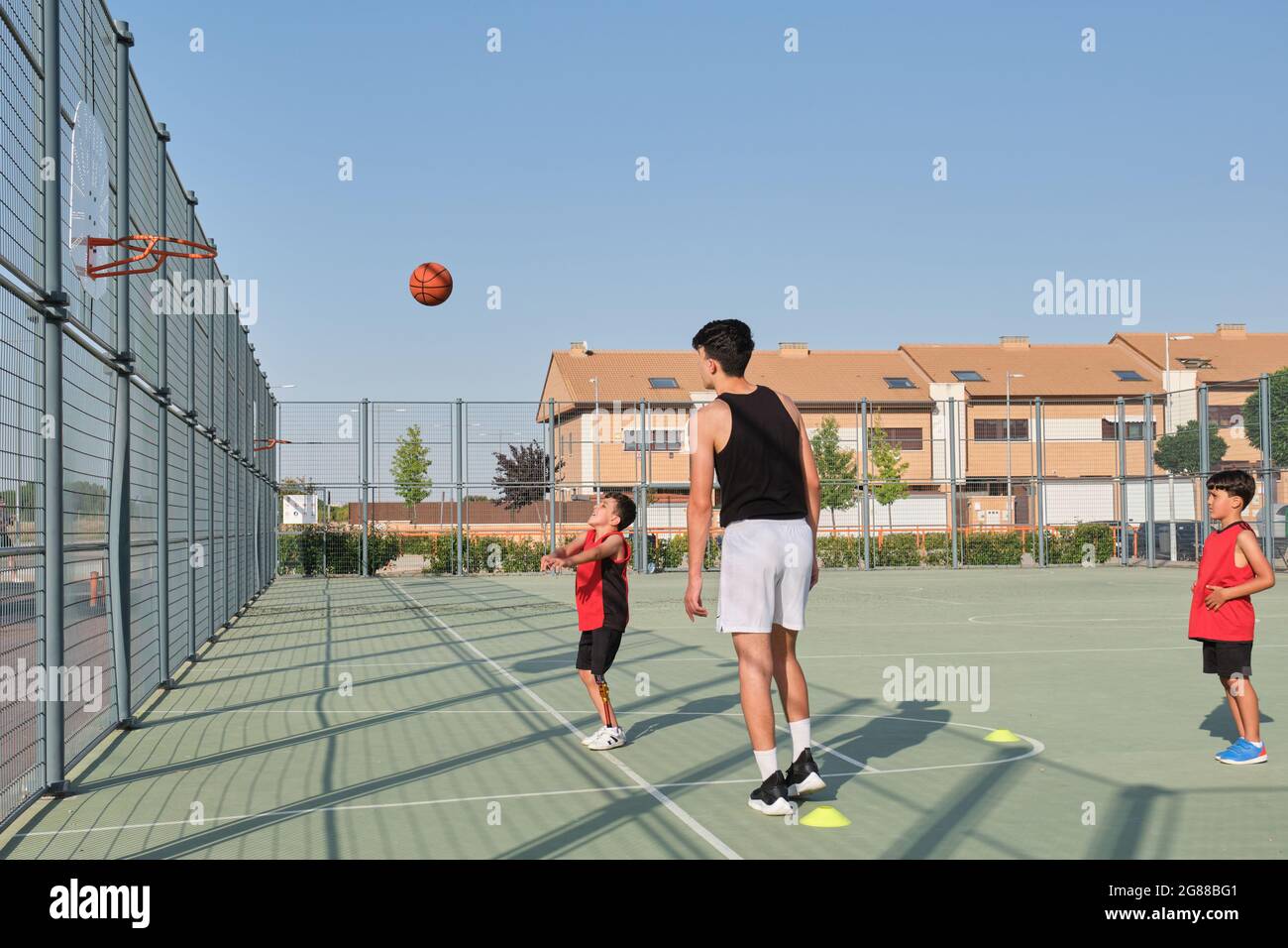 Older brother teaching how to shoot a basketball to his younger siblings, one of them has a leg prosthesis. Playing basket. Stock Photo
