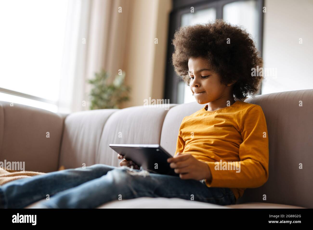 Black curly boy using tablet computer while sitting on sofa at home Stock Photo