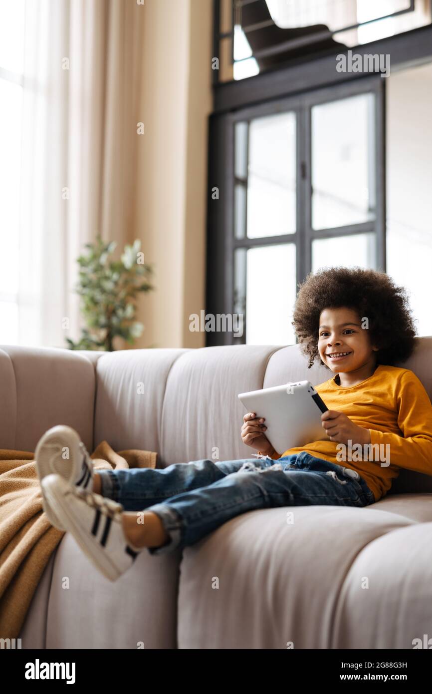 Black curly boy smiling and using tablet computer while sitting on sofa at home Stock Photo