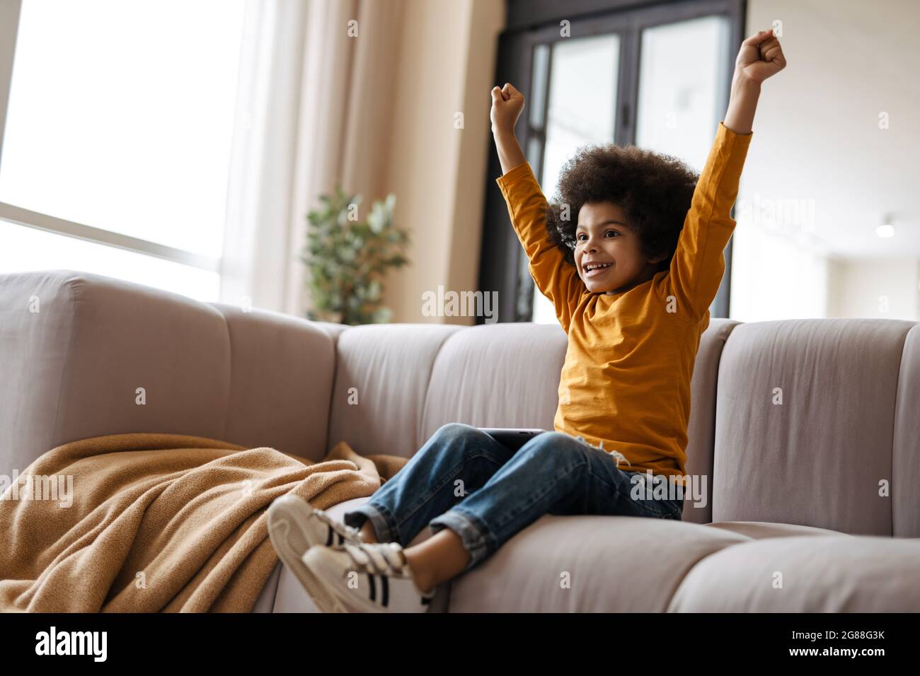 Black curly boy gesturing and using tablet computer while sitting on sofa at home Stock Photo