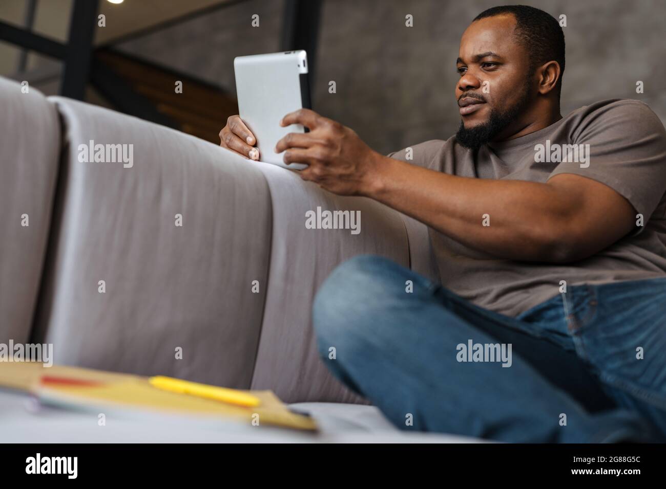 Black unshaven man using tablet computer while sitting on sofa at home Stock Photo
