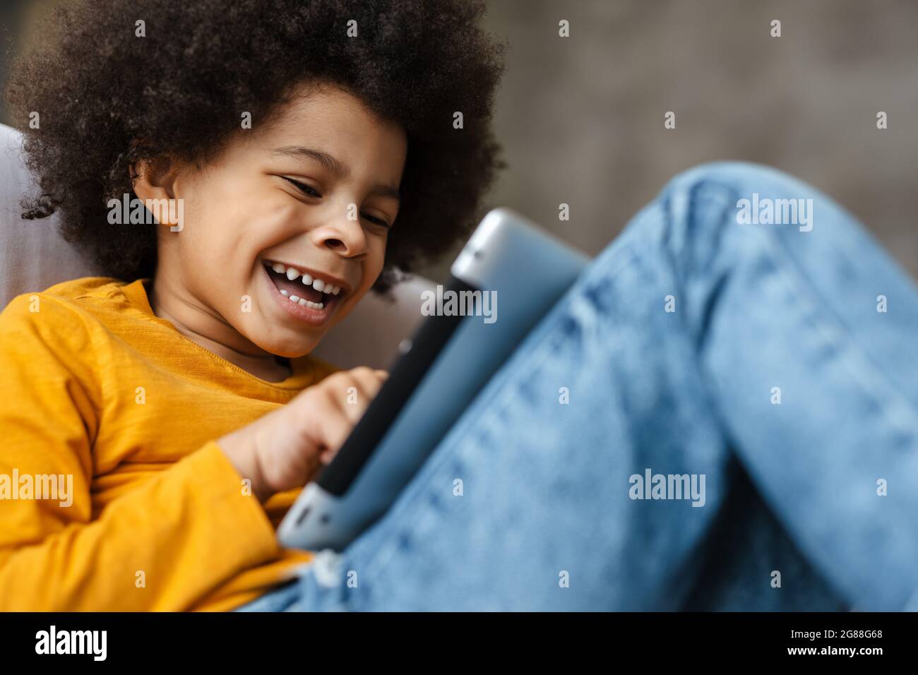 Black curly boy smiling and using tablet computer while sitting on sofa at home Stock Photo