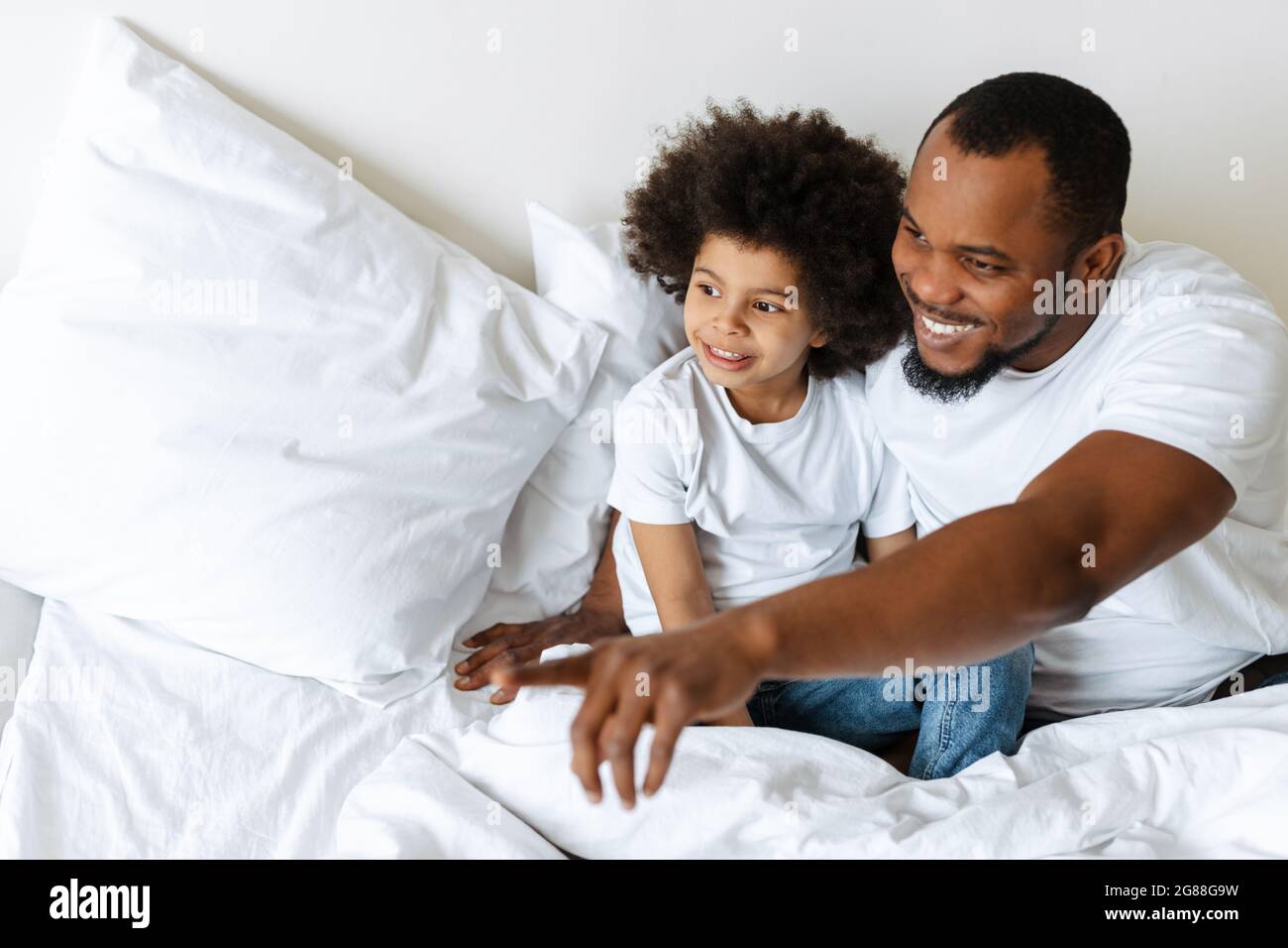 Black father and son smiling and looking aside while sitting in bed at home Stock Photo