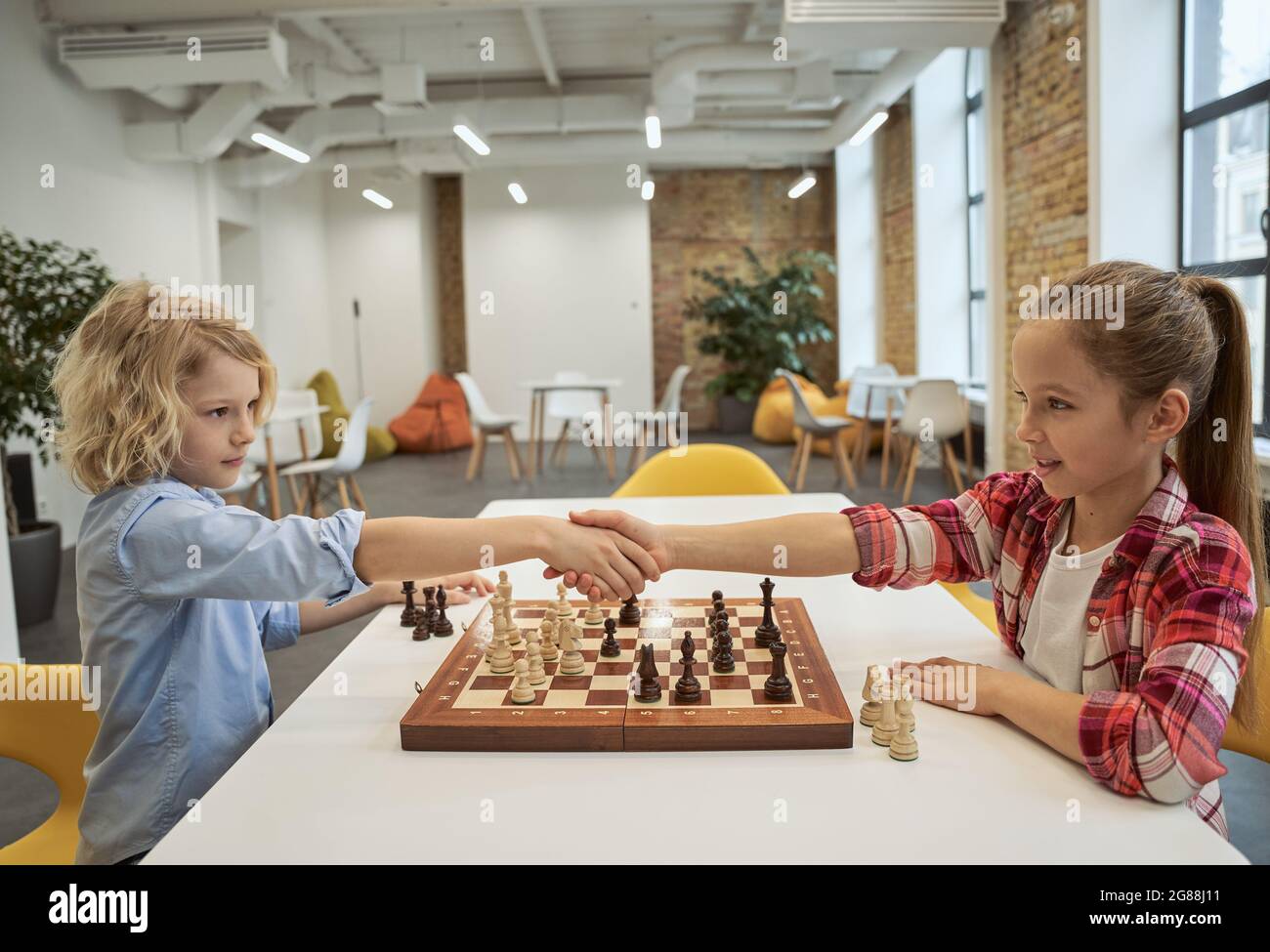 Respect. Adorable little friends, caucasian boy and girl shaking hands after match, playing chess, sitting together at the table in school Stock Photo
