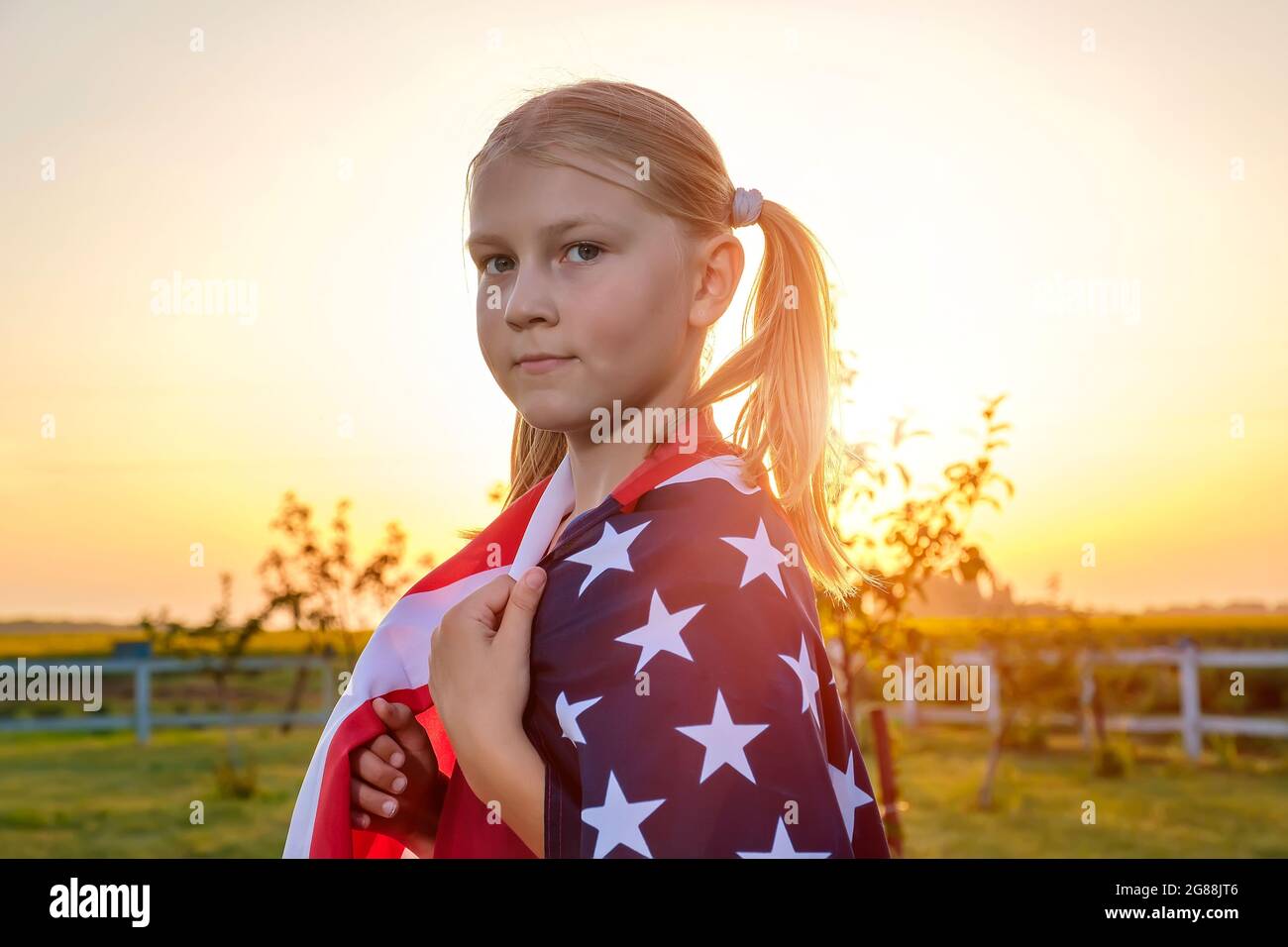 Portrait of a cute little girl, wrapped in an American flag, standing in a field Stock Photo