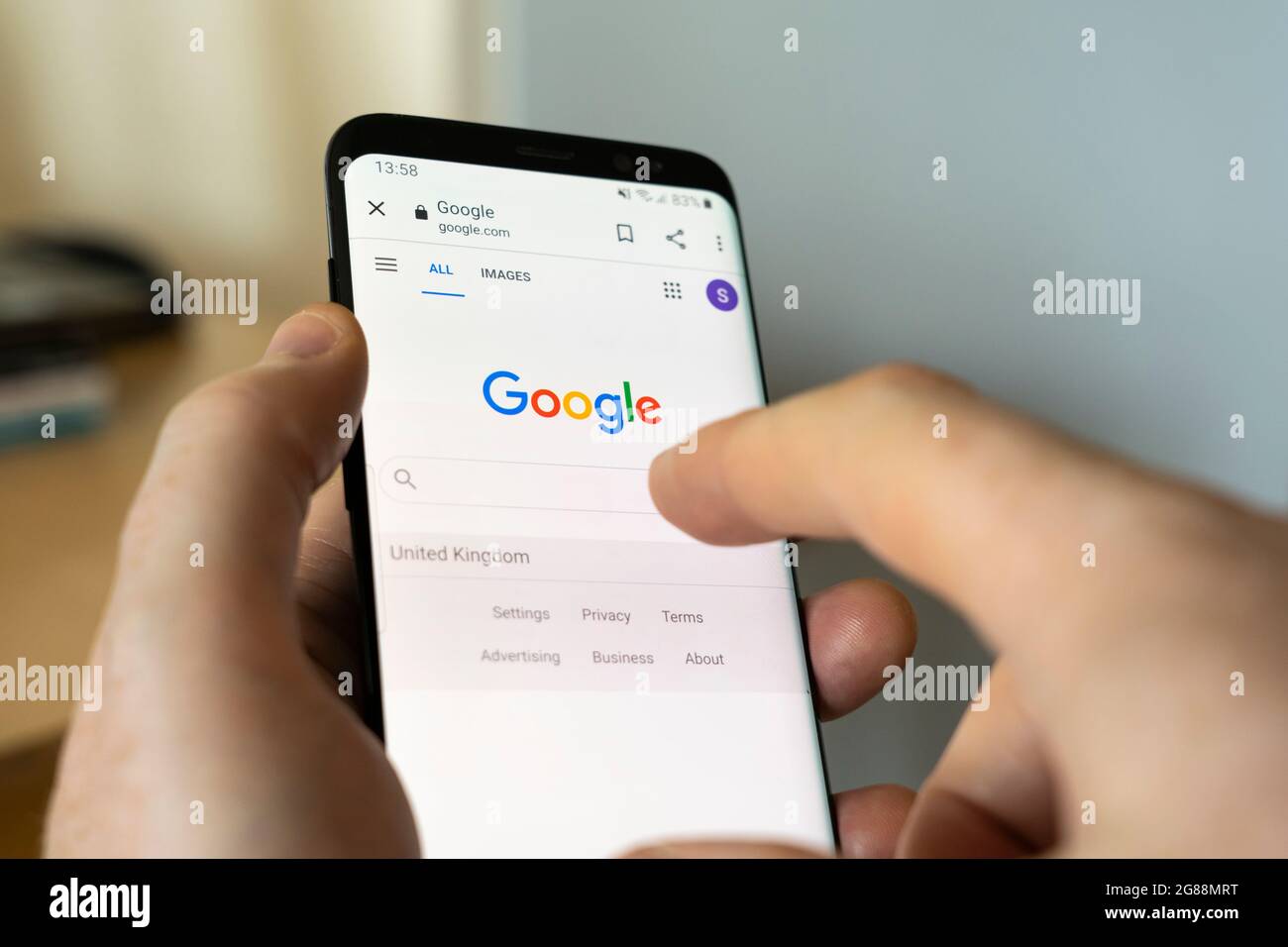A man's hand holding an Android smartphone at home and selecting the Google Search bar on the google.com search engine homepage Stock Photo