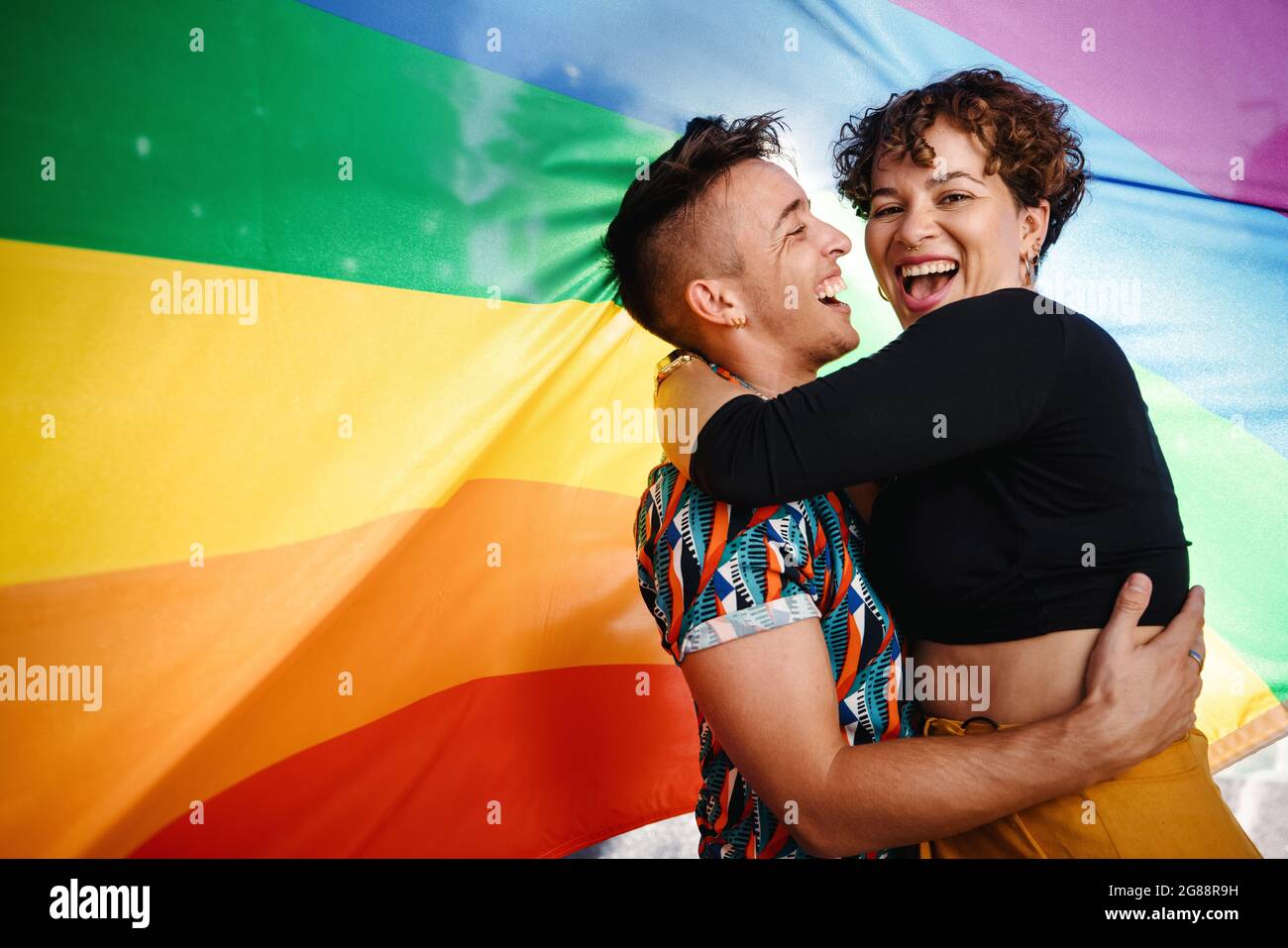 Happy queer couple standing against a rainbow pride flag. Young LGBTQ