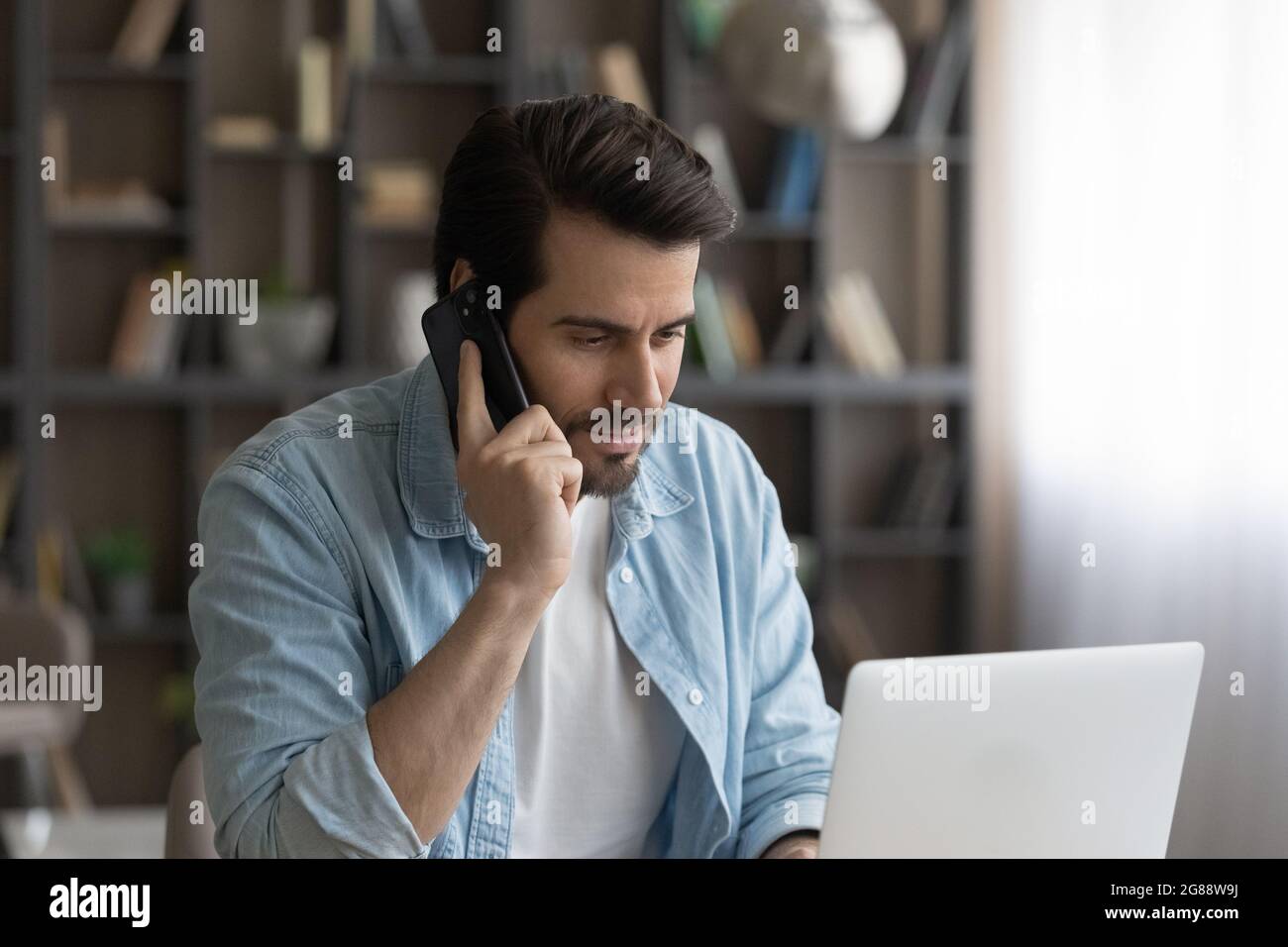Concentrated young man talking on phone, working on computer. Stock Photo