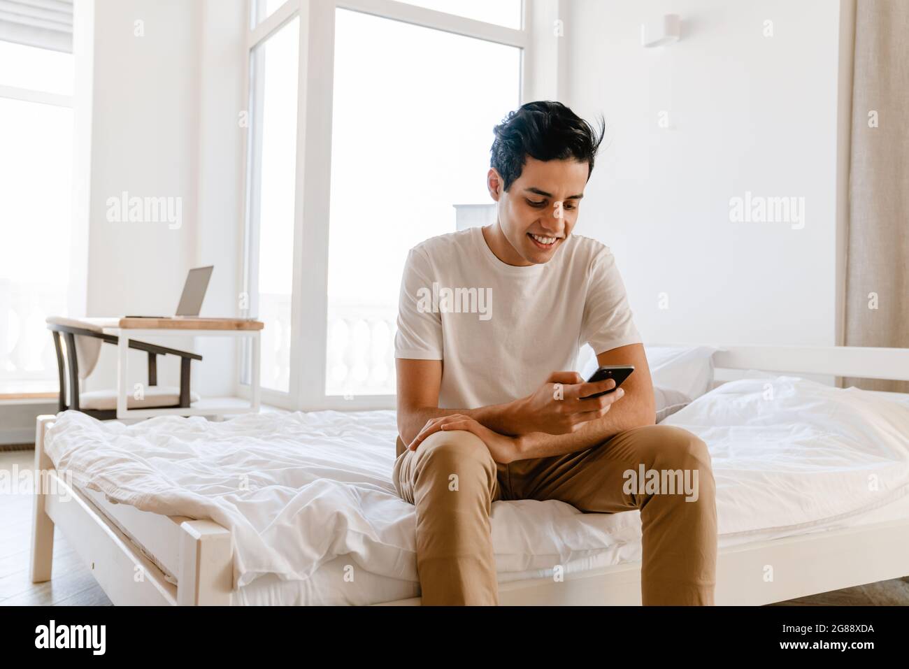 Smiling young hispanic man using mobile phone while sitting on bed at home Stock Photo
