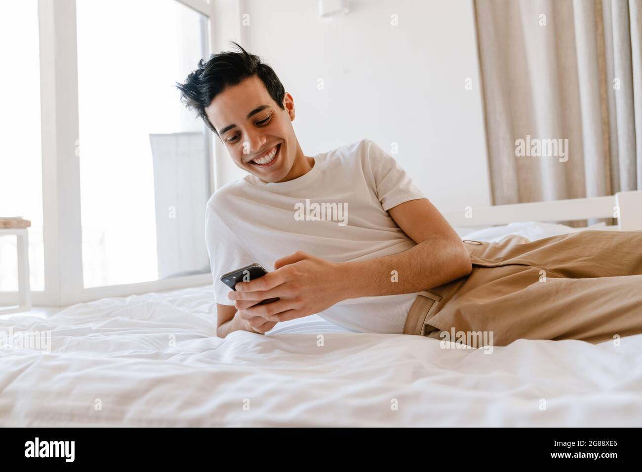 Smiling young hispanic man using mobile phone while laying on bed at home Stock Photo
