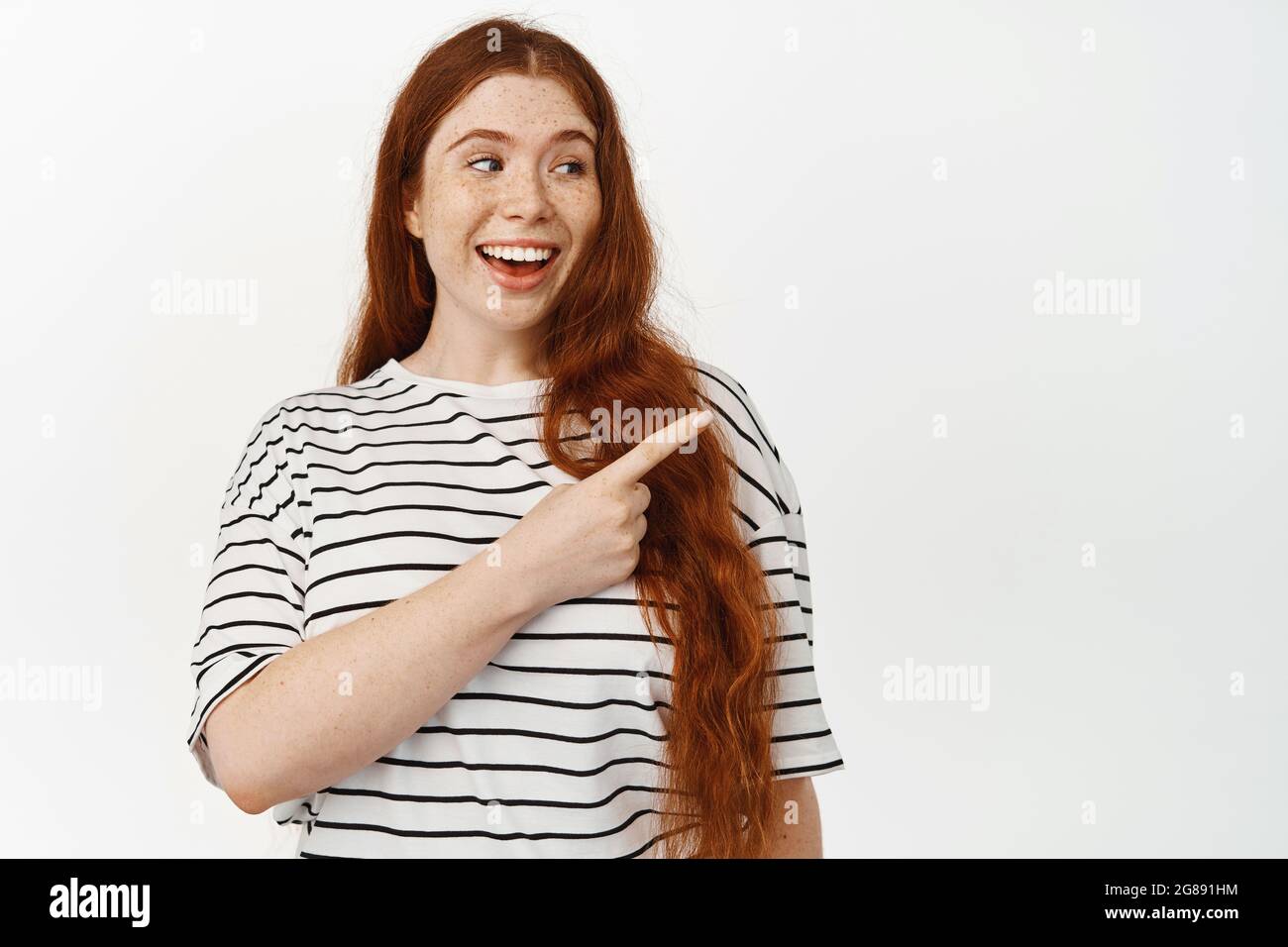 Portrait of pretty teen girl with long red hair, laughing, smiling while pointing finger right at sale banner, showing discount copy space, standing Stock Photo