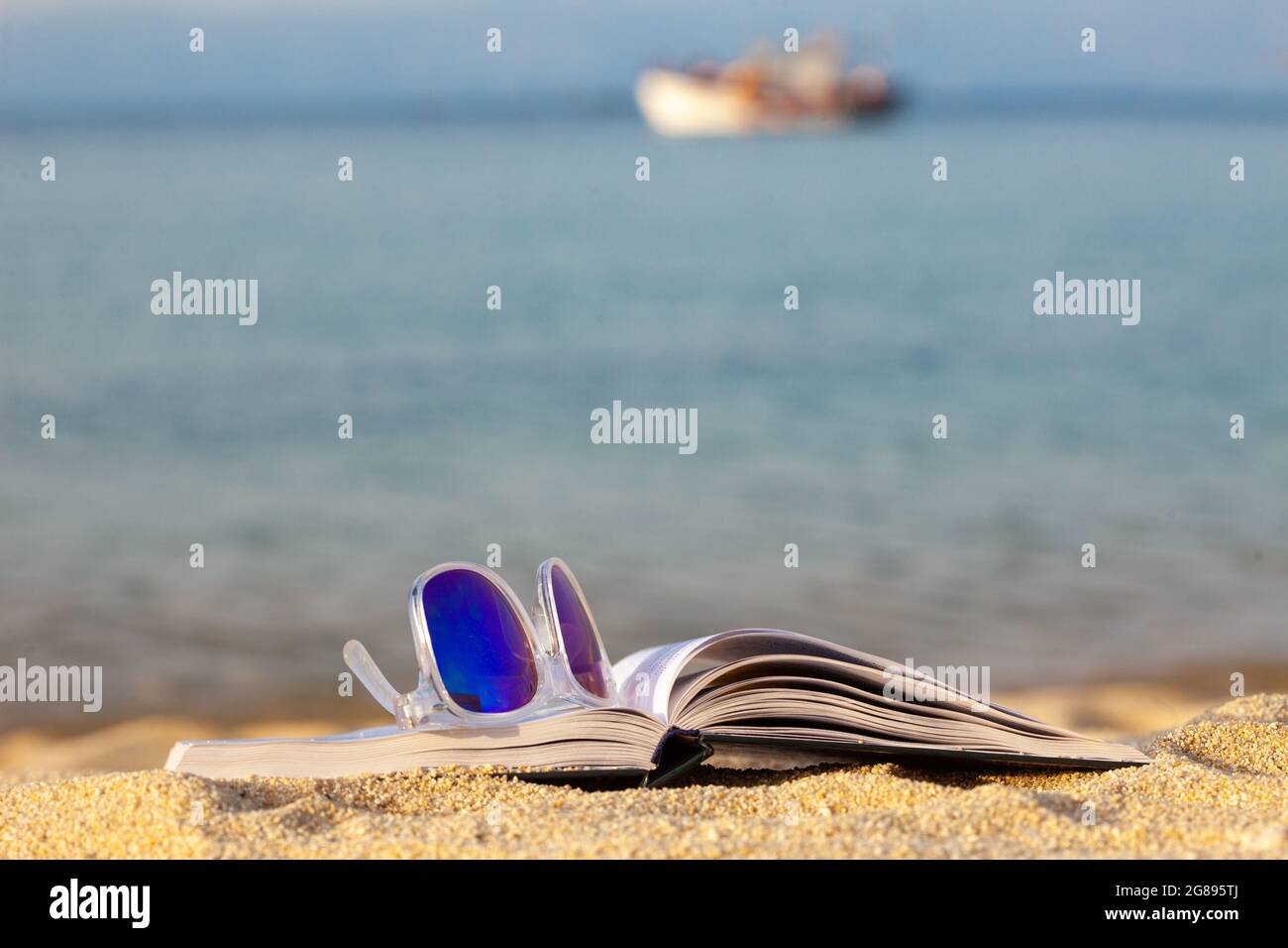 Open book and sun glasses on the beach with small ship in the ...