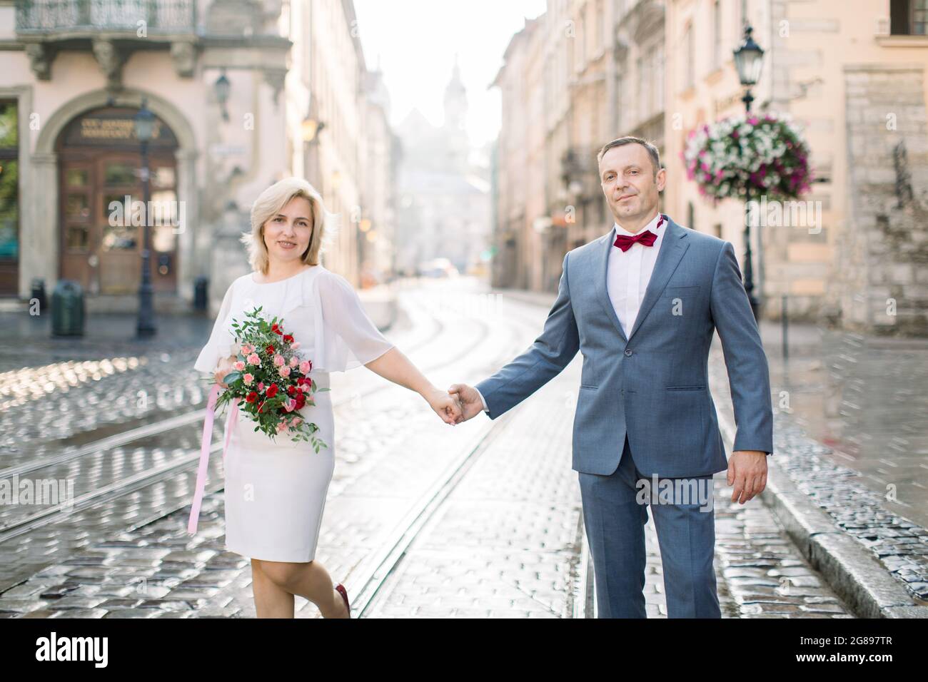 Romantic handsome man and beautiful woman, wearing elegant clothes, enjoying city walk. Portrait of happy middle aged couple in love, holding hands, having a date in ancient city Stock Photo