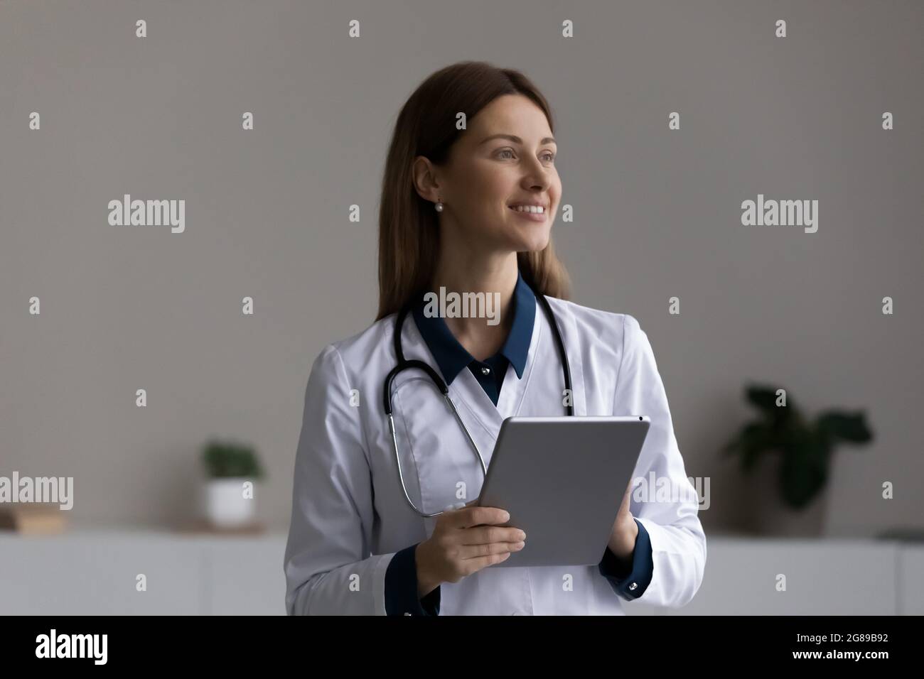 Happy thoughtful female doctor, physician using tablet computer at office Stock Photo