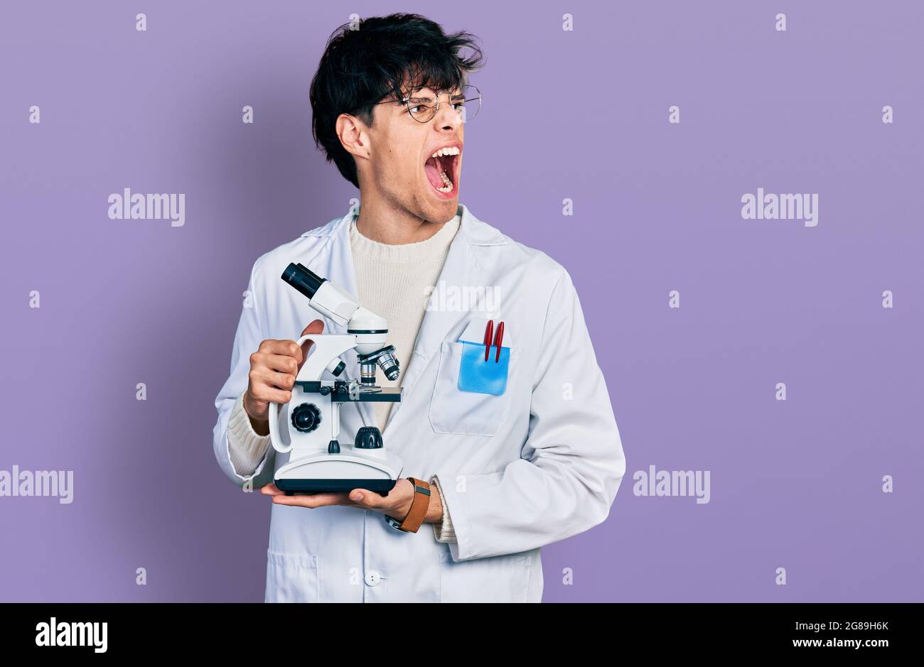 Handsome hipster young man wearing lab coat holding microscope angry ...