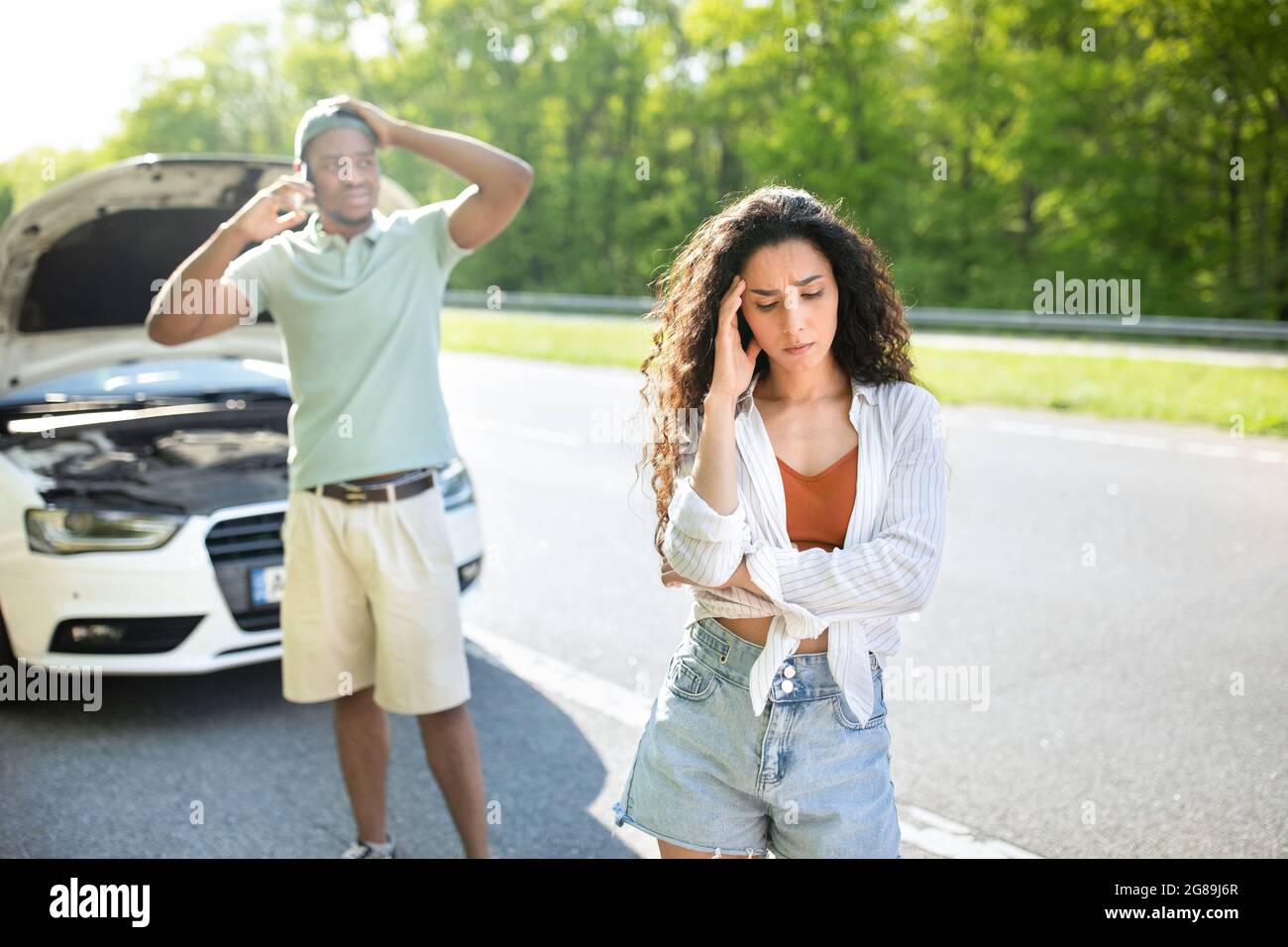Young stressed woman standing on highway near broken car while boyfriend calling breakdown road service on smartphone Stock Photo