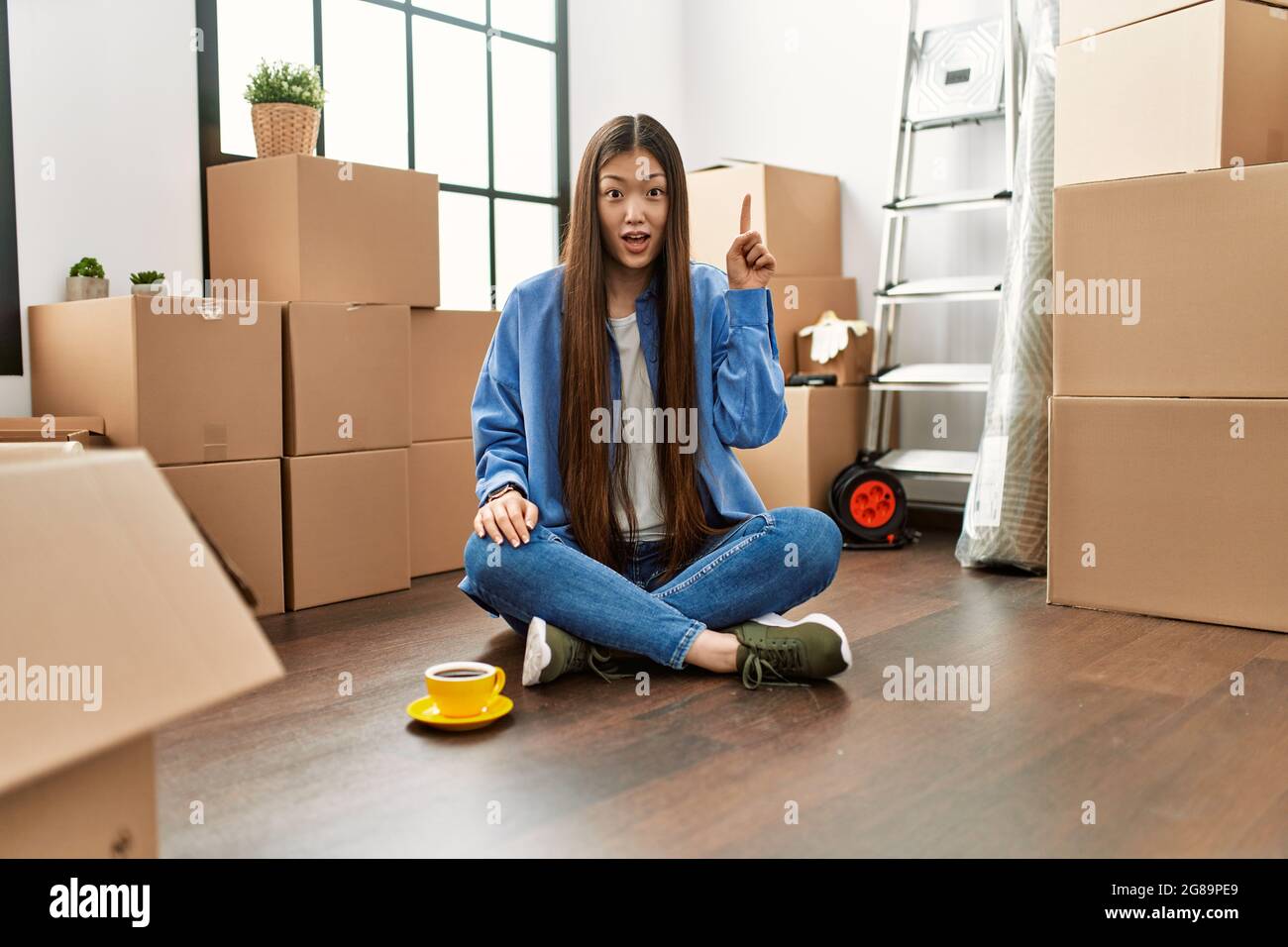 Young chinese girl sitting on the floor at new home pointing finger up ...