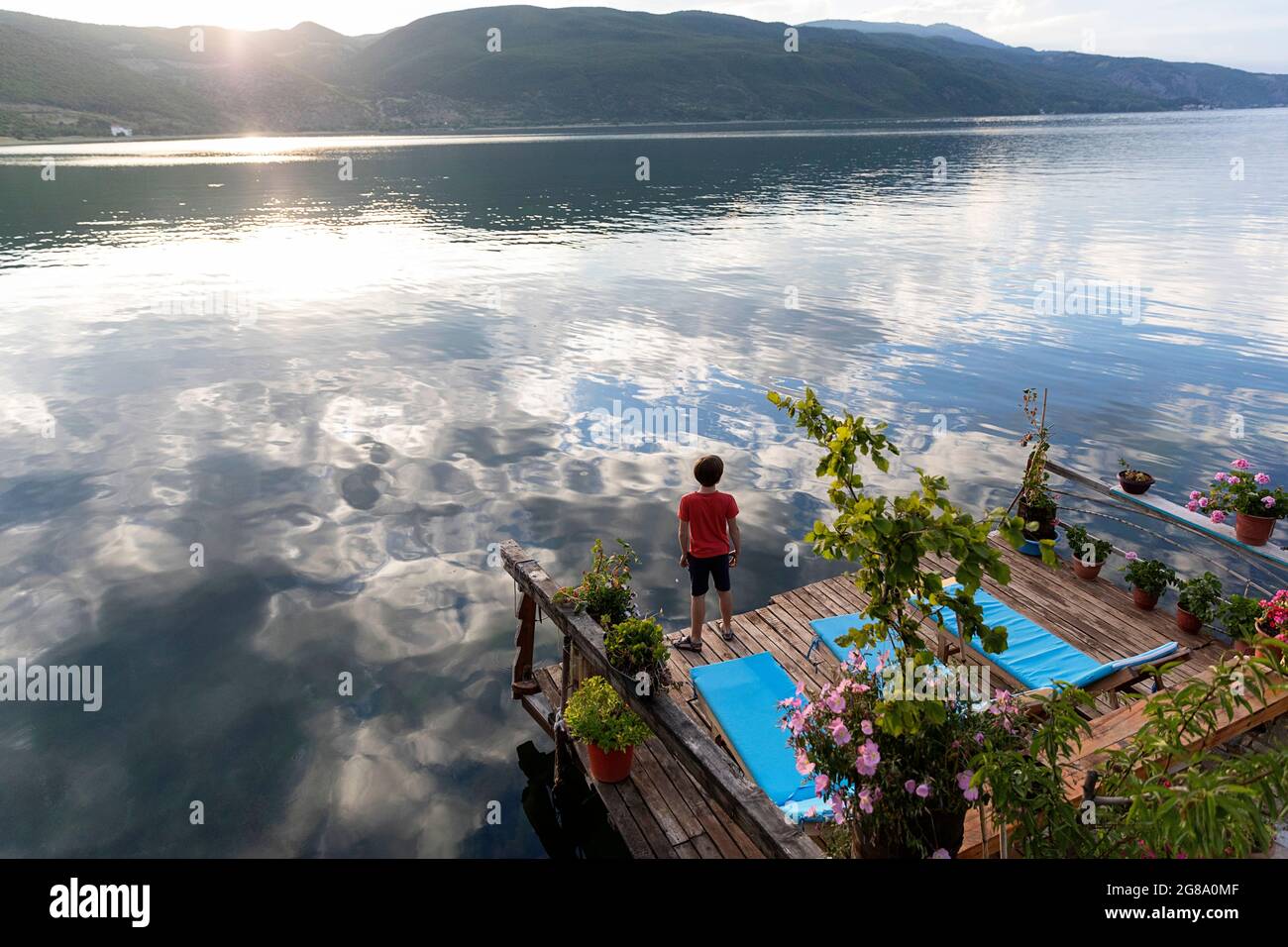 Boy standing by wooden lounge chairs at sunset on a pier on the shores of the calm lake Ohrid, Lin, Albania Stock Photo