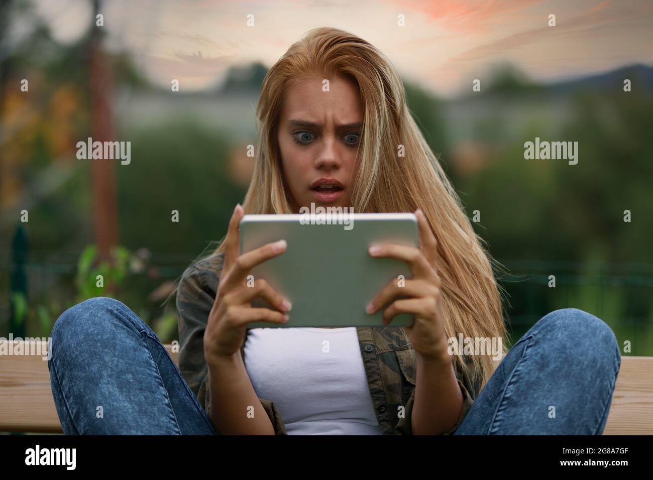 Horrified young woman staring wide-eyed at her tablet with mouth open and a flabbergasted expression as she sits with knees up on an outdoor bench Stock Photo