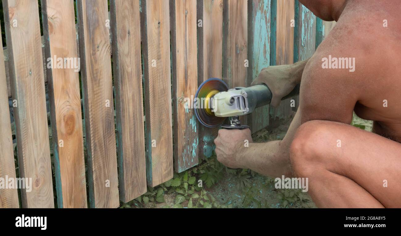 A man cleans a wooden fence from paint with a small sander. Stock Photo