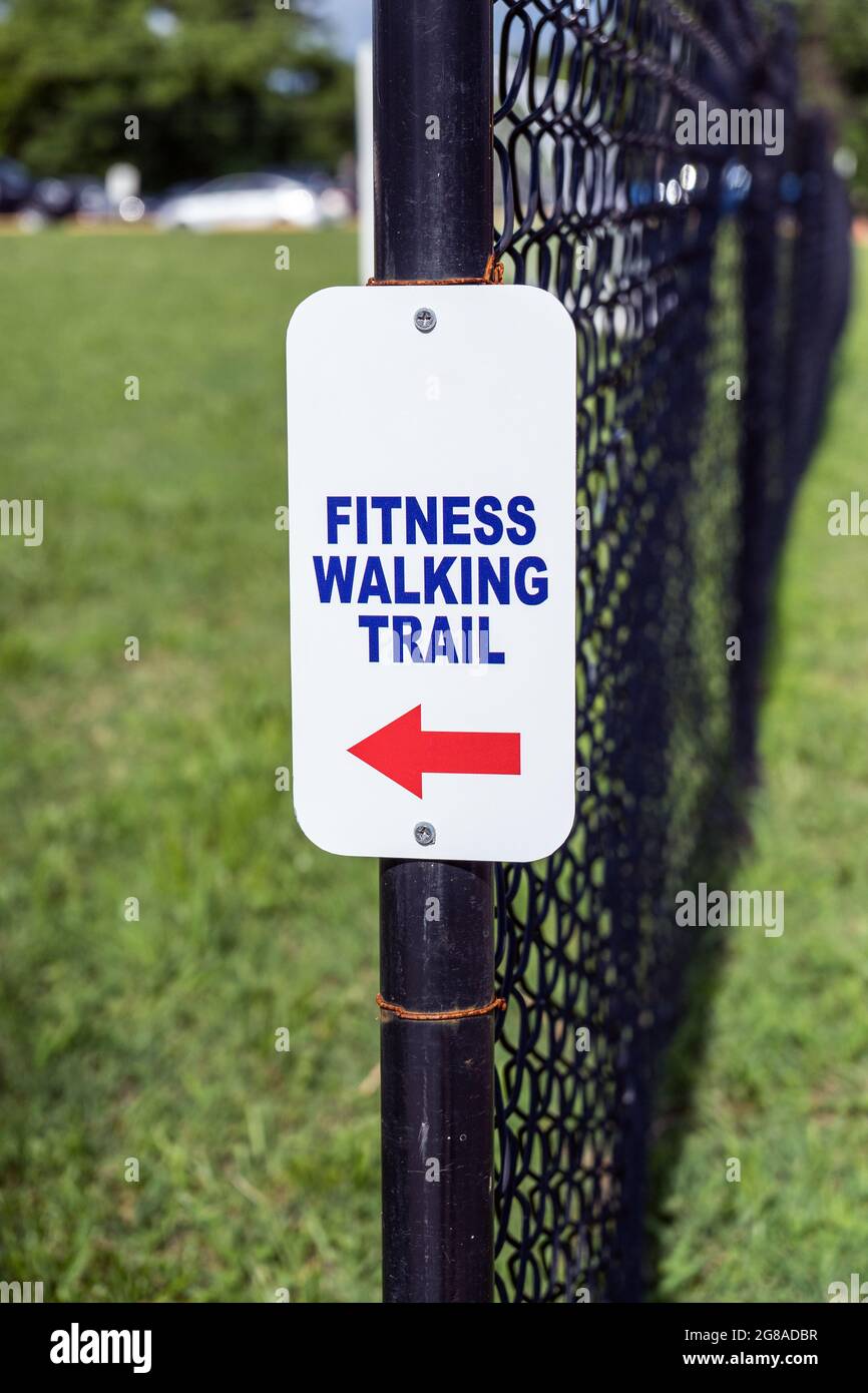 white sign for a fitness walking trail with blue letters and red arrow mounted on a black chain link fence surrounded by green grass Stock Photo