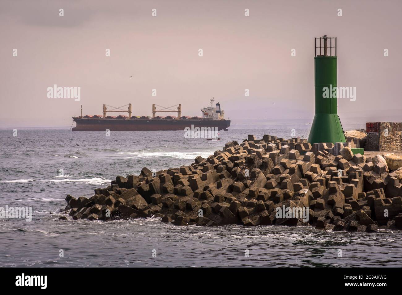 Breakwater made with concrete cubes and a green tower with a little boat and a big ship in the sea. Antofagasta, Chile Stock Photo