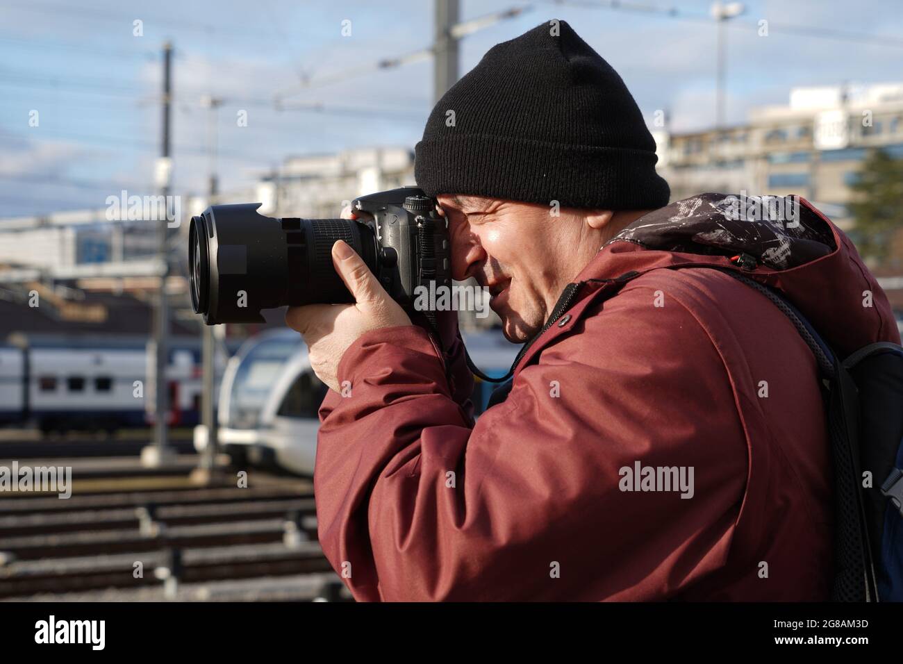 A man taking photos of trains in winter. Side view photo. He is wearing a cap and winter jacket. Stock Photo