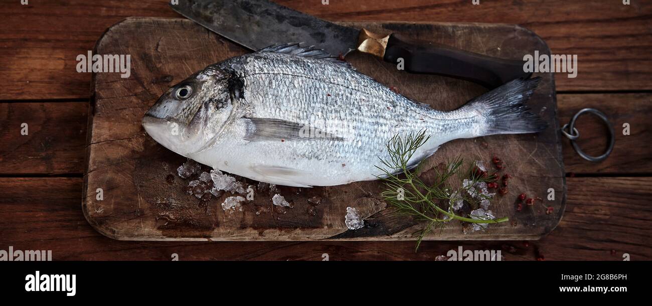 Whole raw fresh sea bream on a rustic vintage chopping board with cleaver and sprig of dill in an overhead view in a wide angle panorama banner or hea Stock Photo