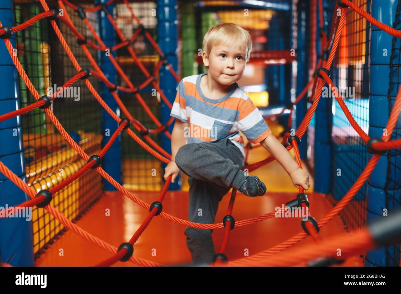 Happy boy playing in indoor playground on net trail. Child having fun on modern playground. Cute kid playing on colorful playground at shopping mall Stock Photo