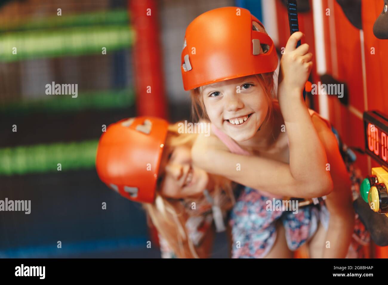 Two happy little girls in red helmets climbing the wall in bouldering center. School girls smiling at the camera and having fun in indoor playground f Stock Photo